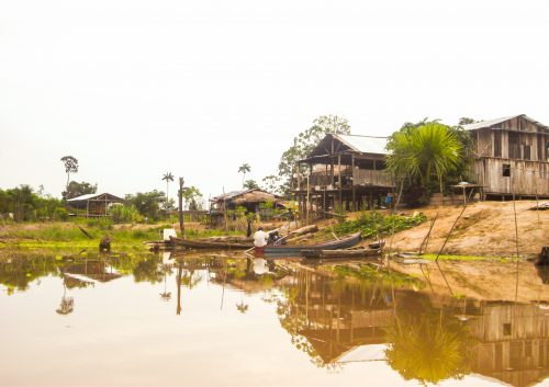 A man on a small canoe can be seen in the river in front of a house in the Amazon jungle.