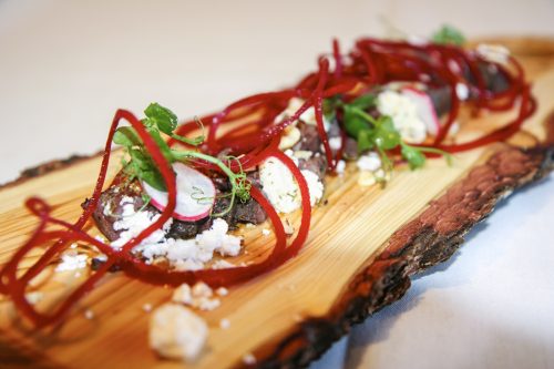 A wooden board displays a colorful dish including cheese, radishes and beetroot.