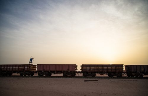 A traveler armed with his surfboard jumps from one train car to another during a beautiful sunset over the Sahara desert.