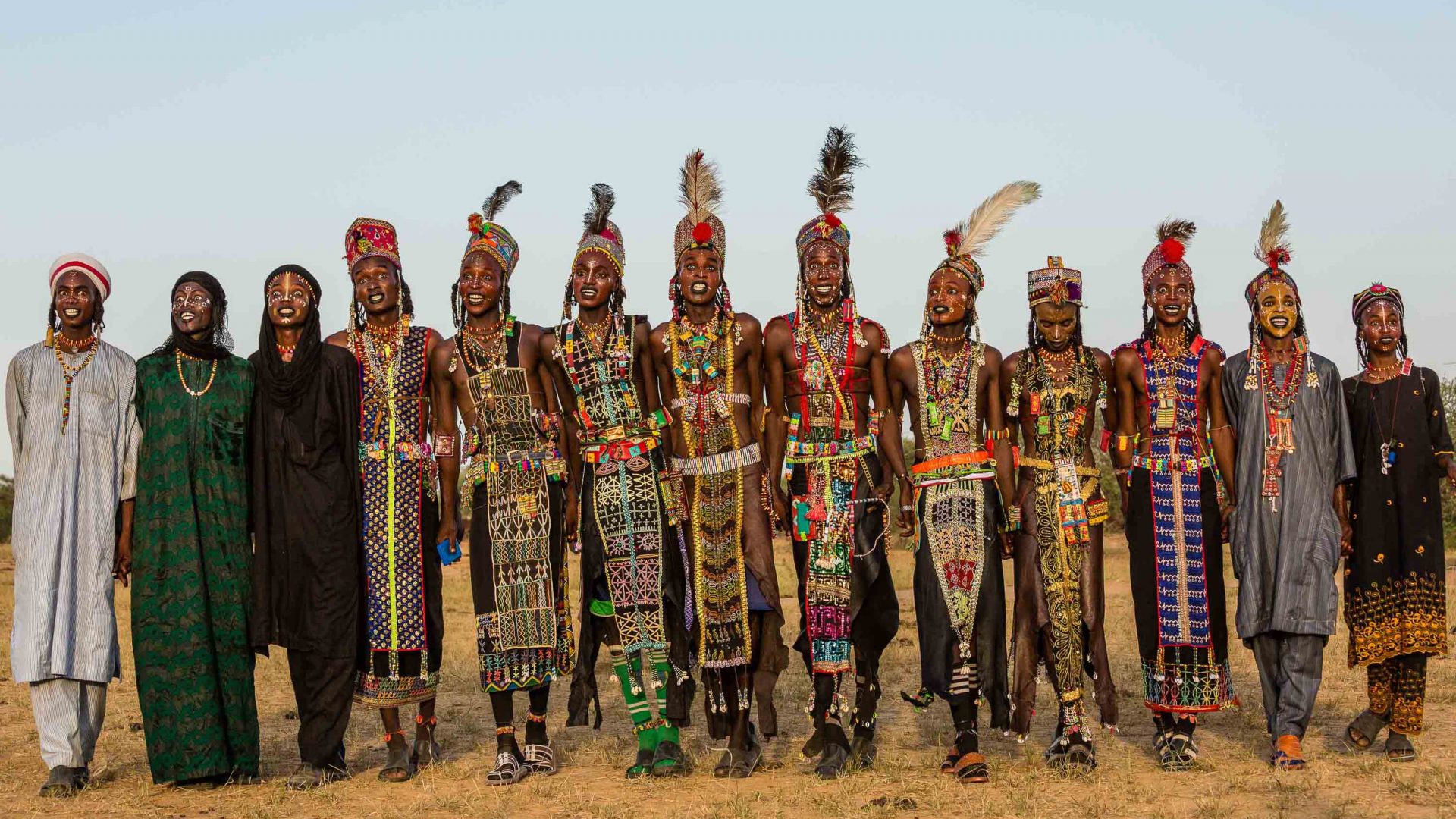 A long line of Wodaabe men and boys, wearing bejeweled leather tunics and sparkling crowns and feathers.