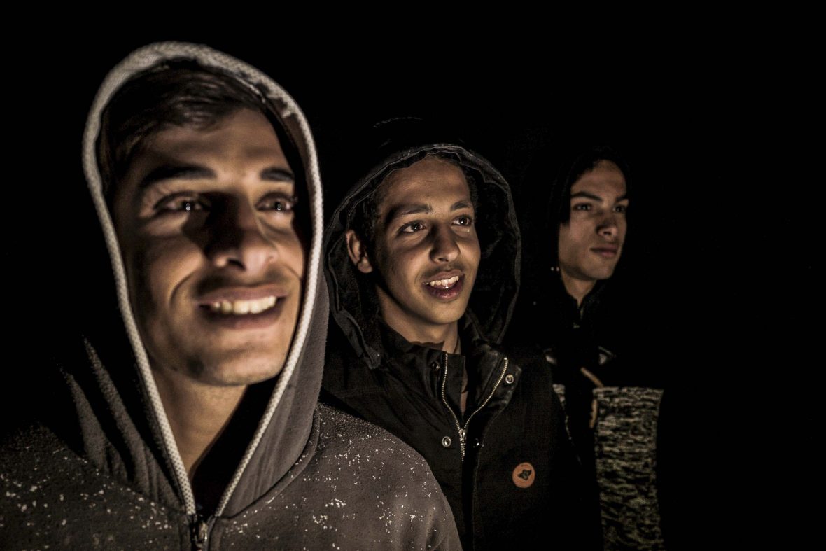 In the darkness, the faces of three young men are illuminated.