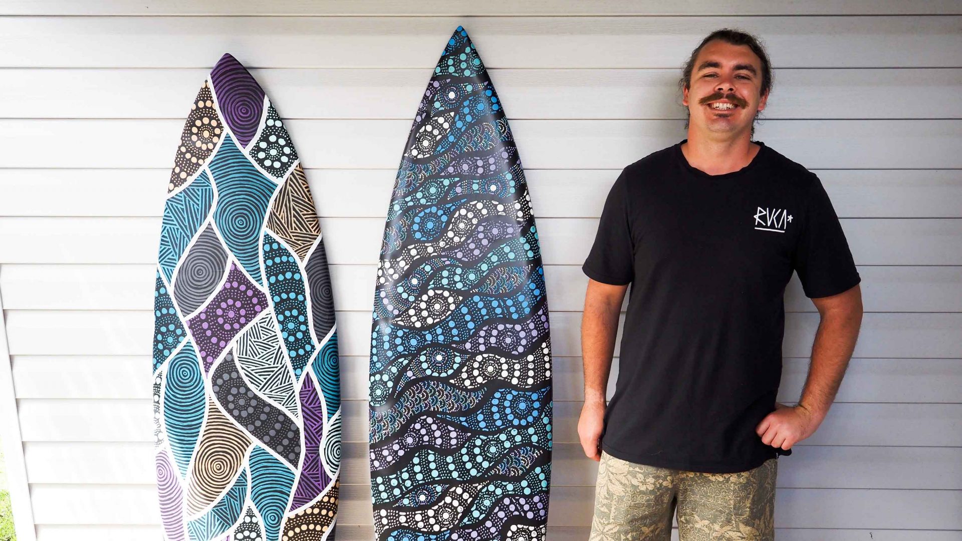Portrait of Zachary standing alongside his painted surfboards.