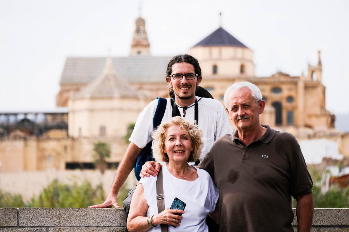 An older couple and a young man smile for the camera in front of an old building.