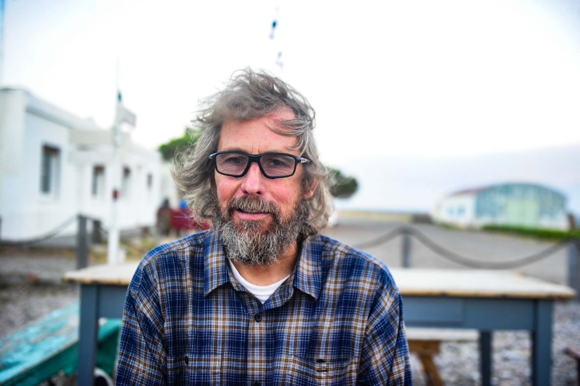 A portrait of Matias wearing a plaid shirt and black glasses. He has a grey beard and grey hair.