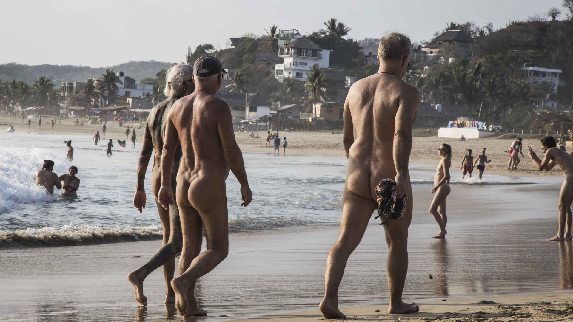 A group of naked men walk down the beach.