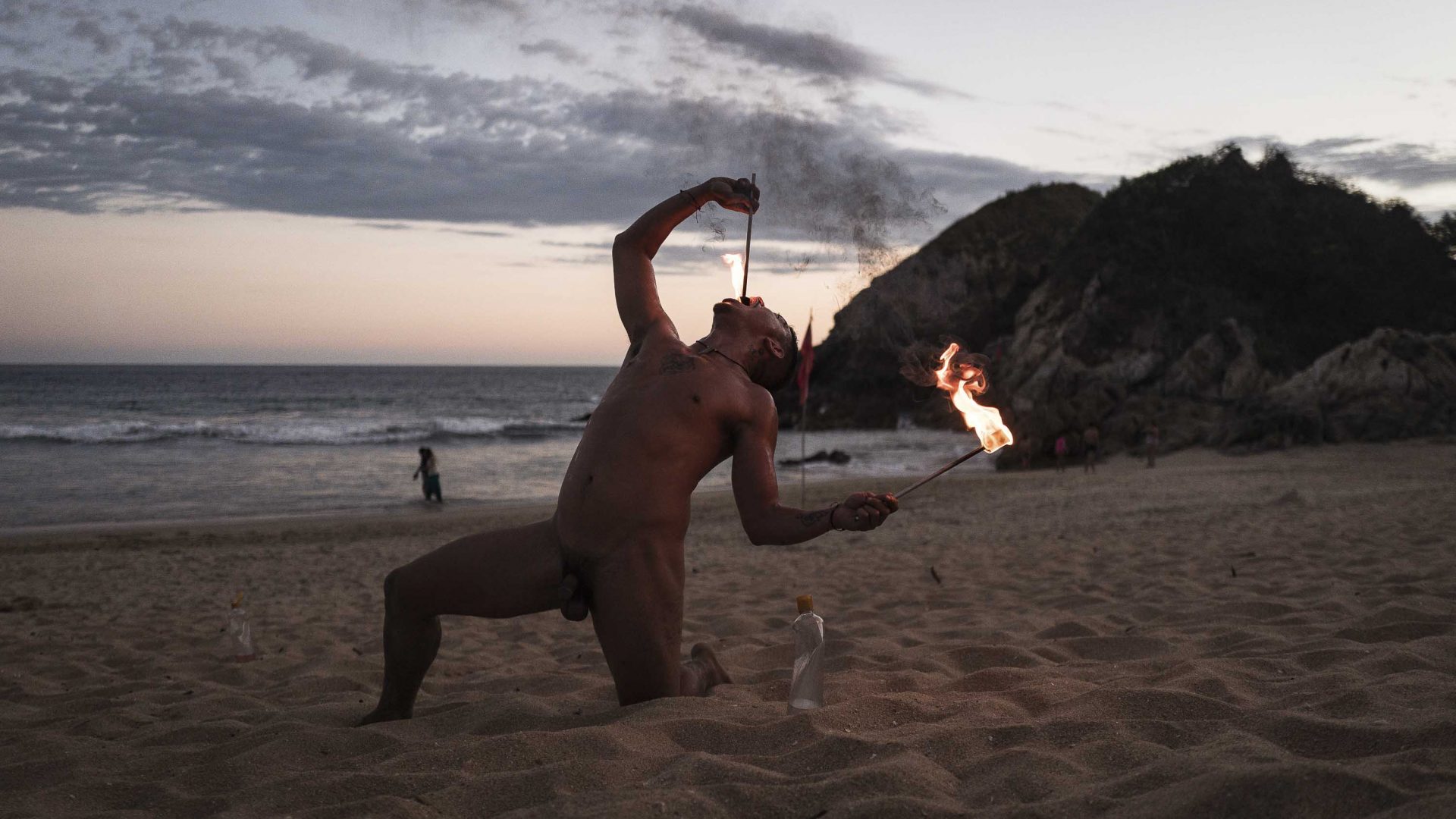 A naked man swallows fire on a beach.