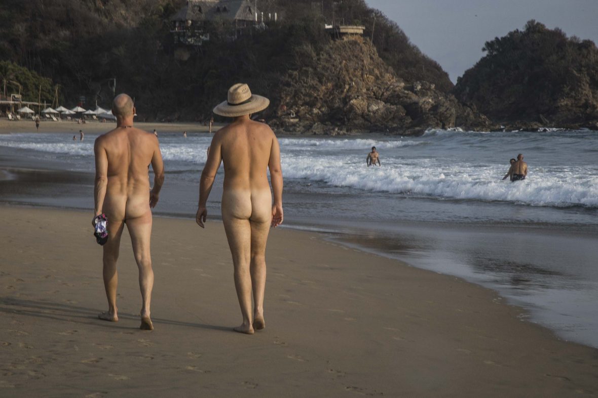 Two naked men walk down a beach.