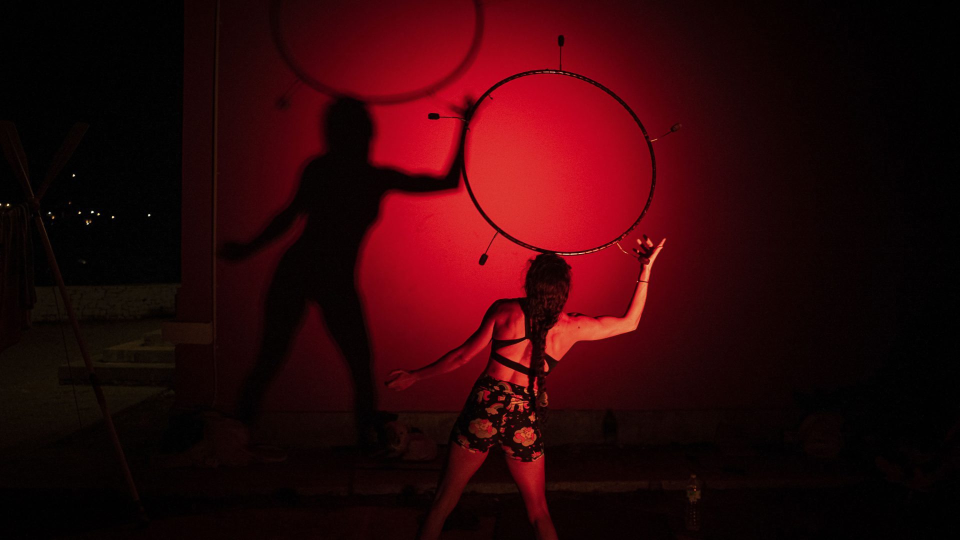 A performer plays with their hula hoop, casting shadows on a red wall.