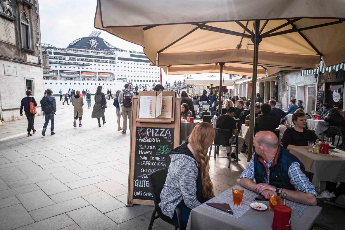 Tourists at a restaurant in Venice watch a cruise ship come in.