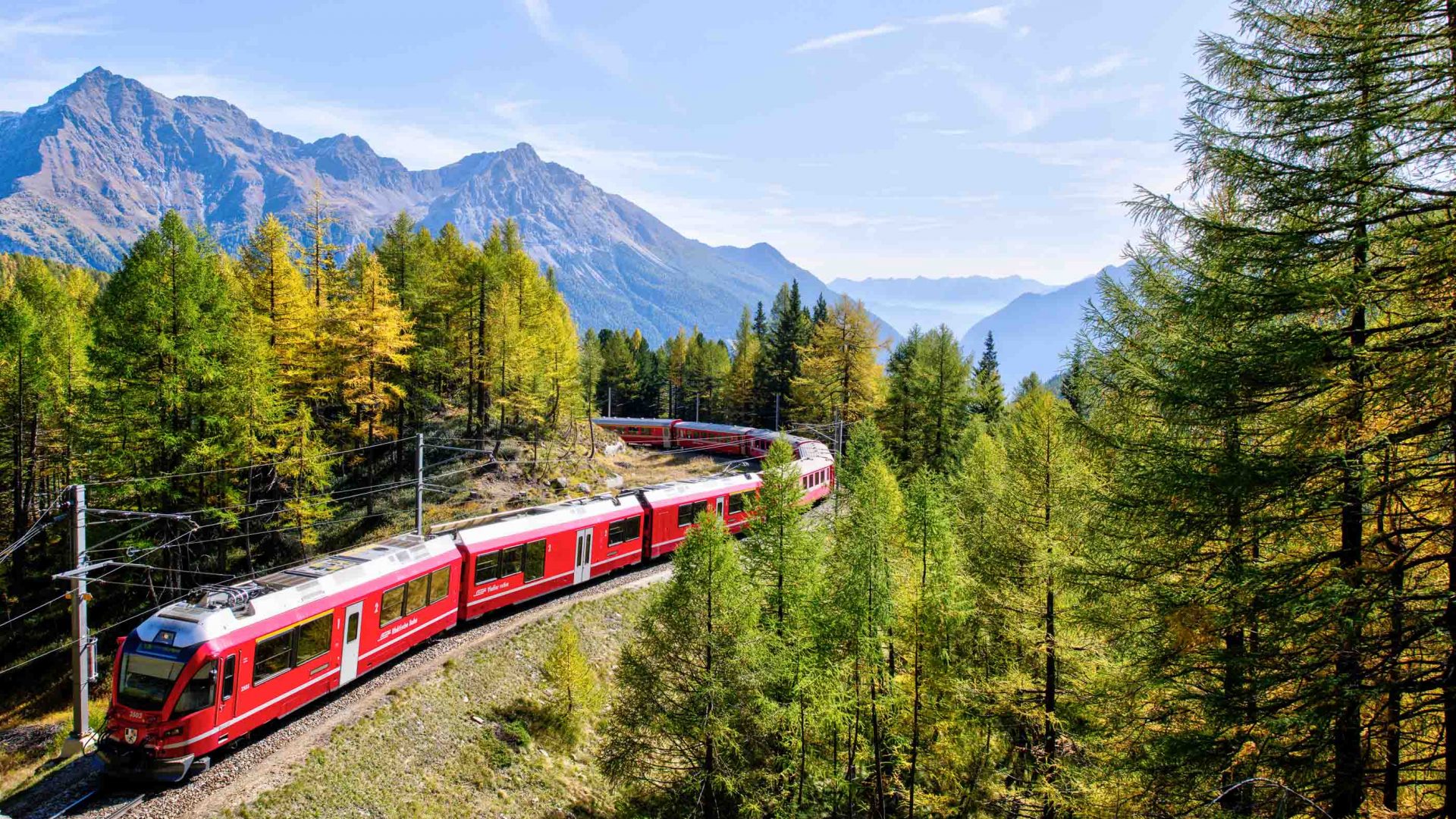 A red train traveling through green trees with mountains in the background.