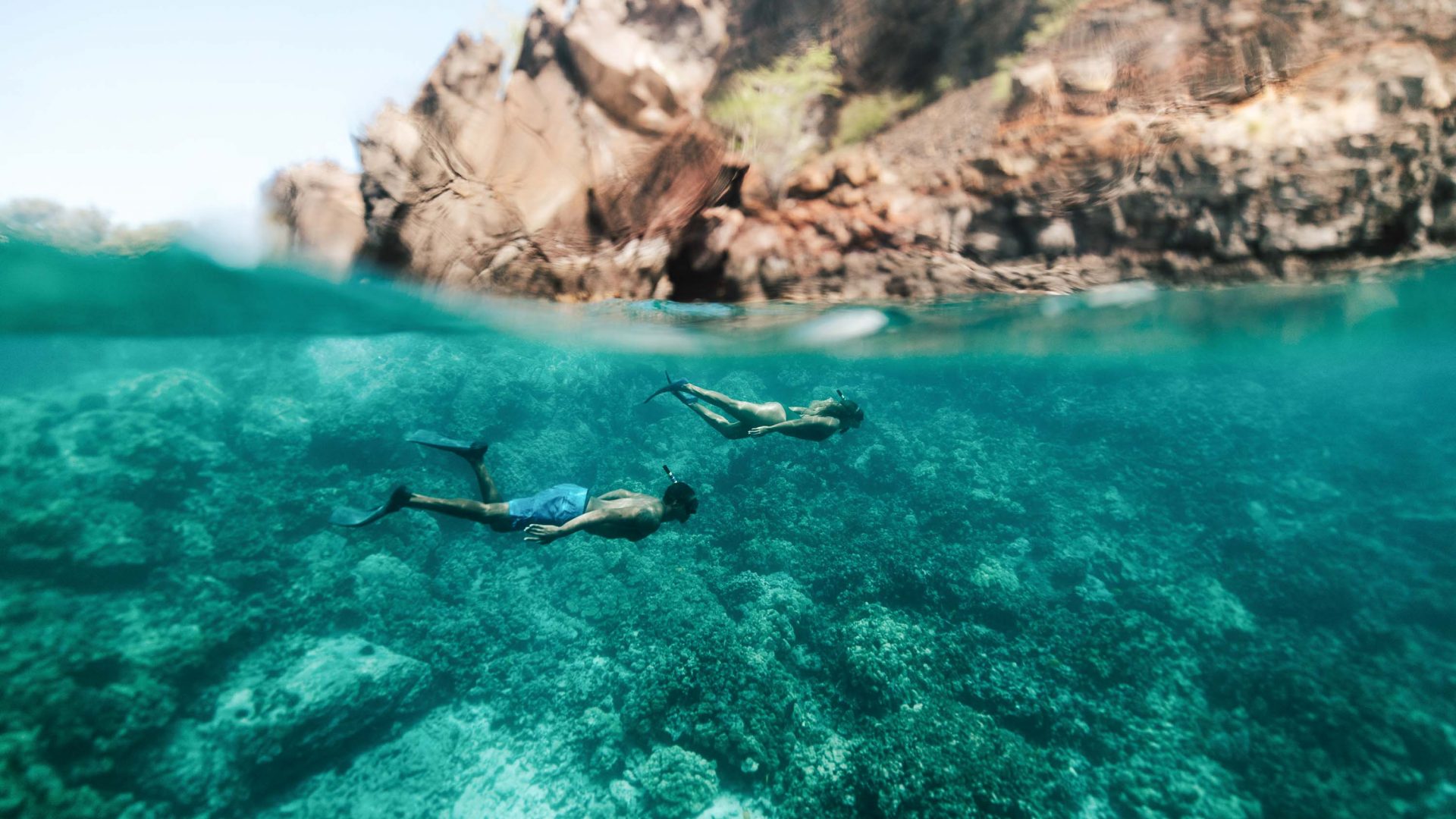 Snorkelers seen just below the surface of the water. Rocks can be seen above them.