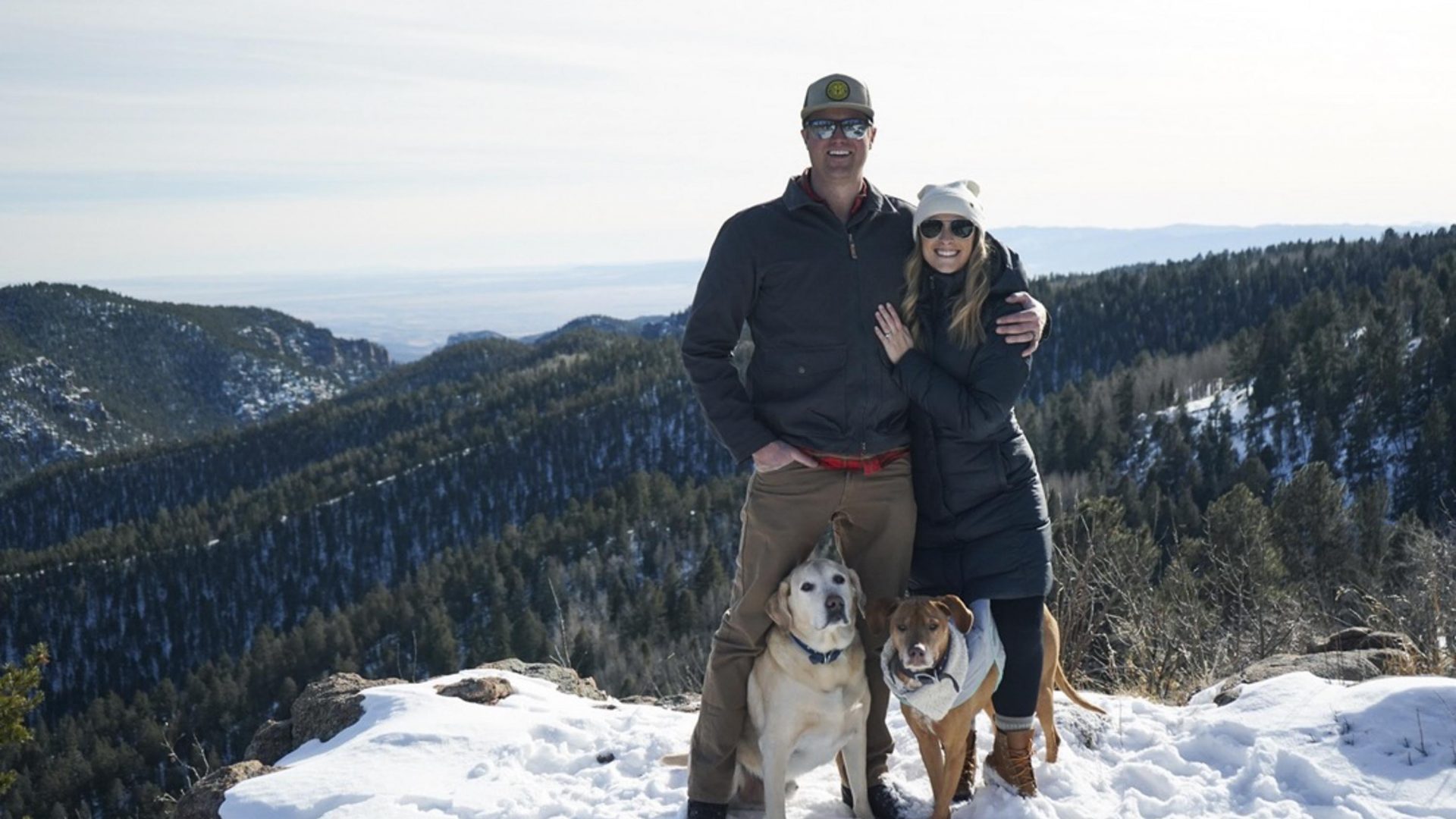 A man and a woman stand on a snowy mountain, backed by trees, with their two dogs.