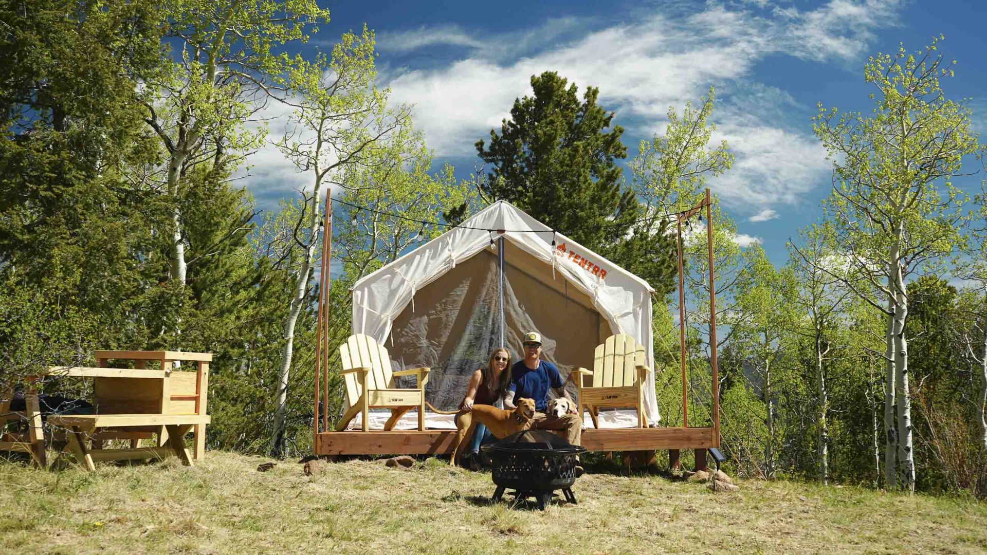 A man and a woman sit together on the porch of a tent which is surrounded by trees.