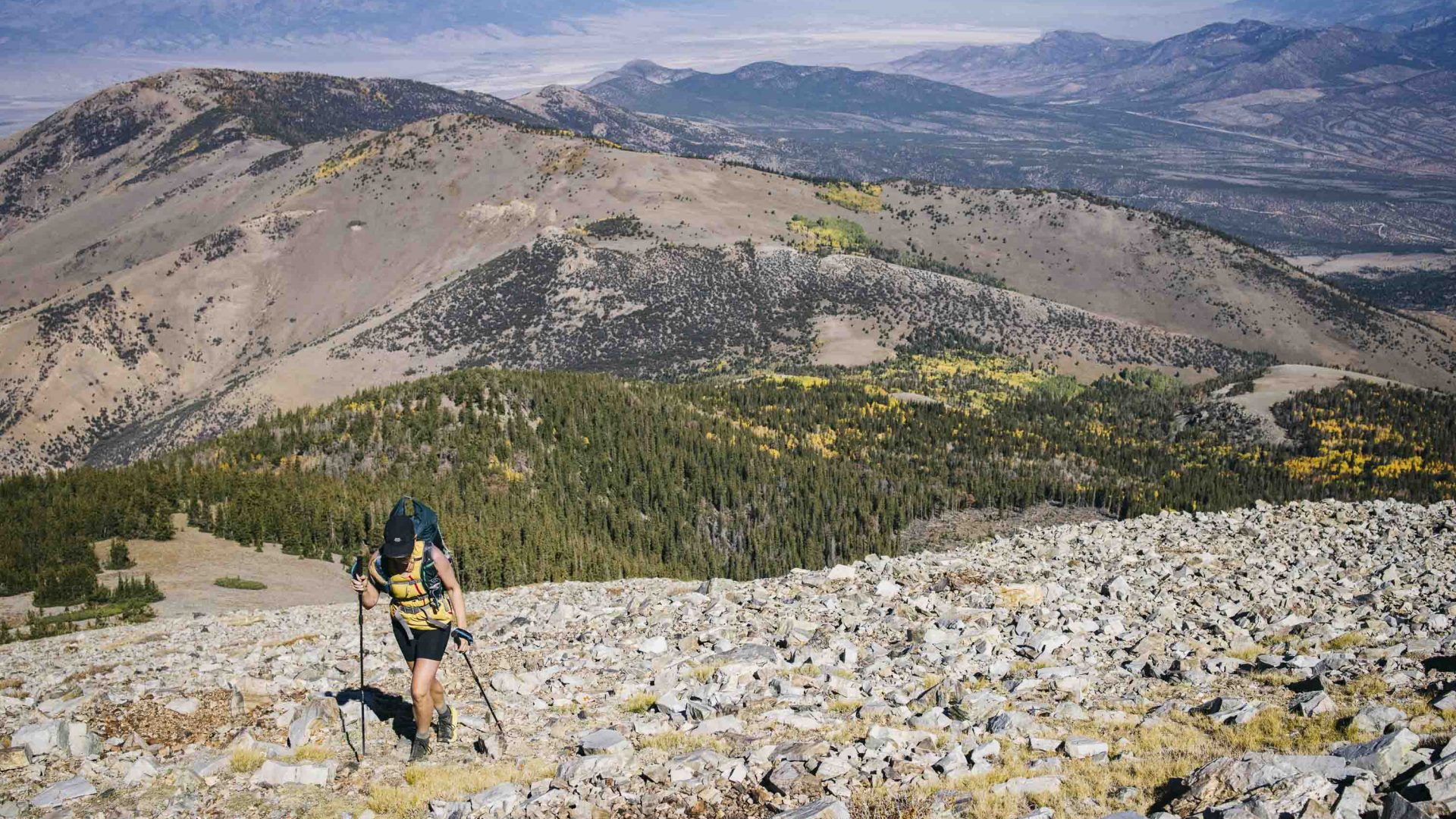 A woman hikes up a rocky hill with trees in the background.