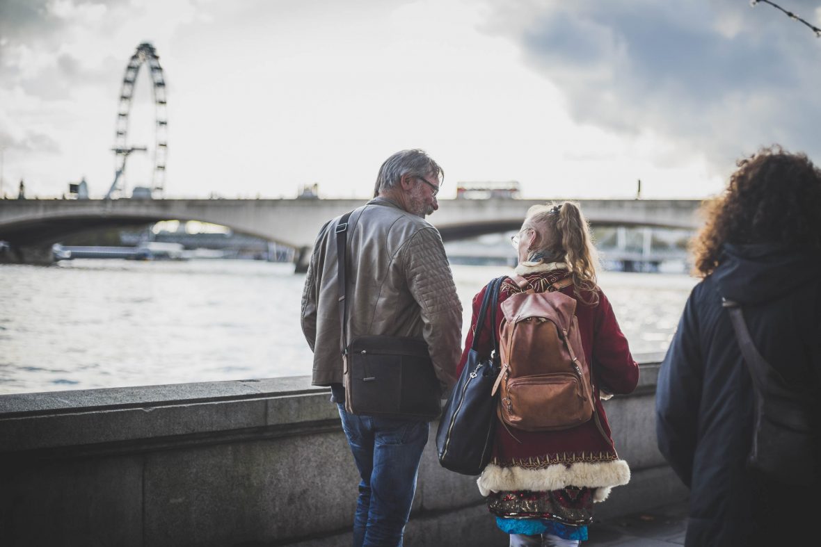 A woman wearing a backpack walks with a man along the river. The London Eye is in the background. We can only see their backs.