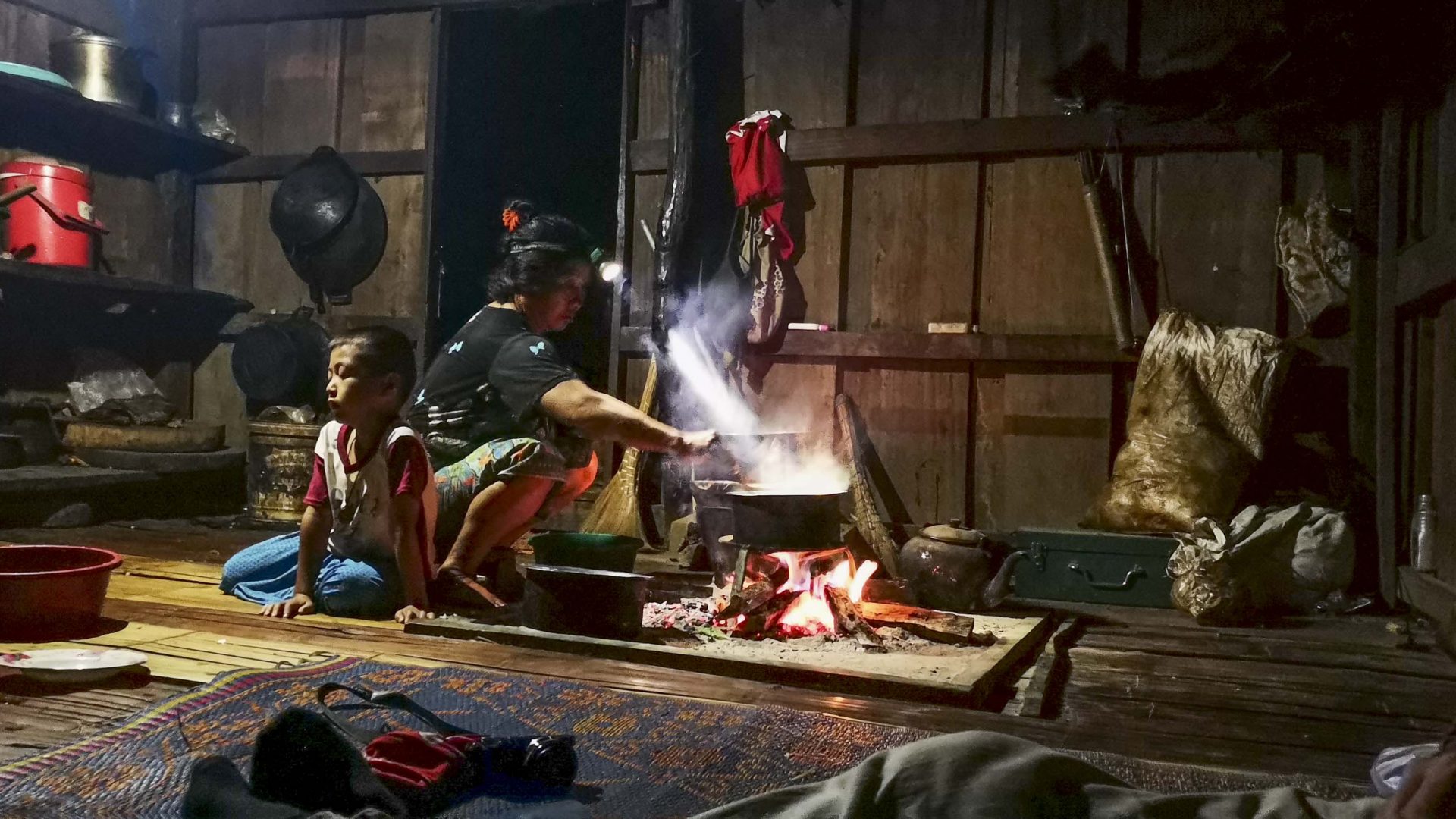 A woman cooks on a fire on the floor in a dimly lit room. A child is by her side.