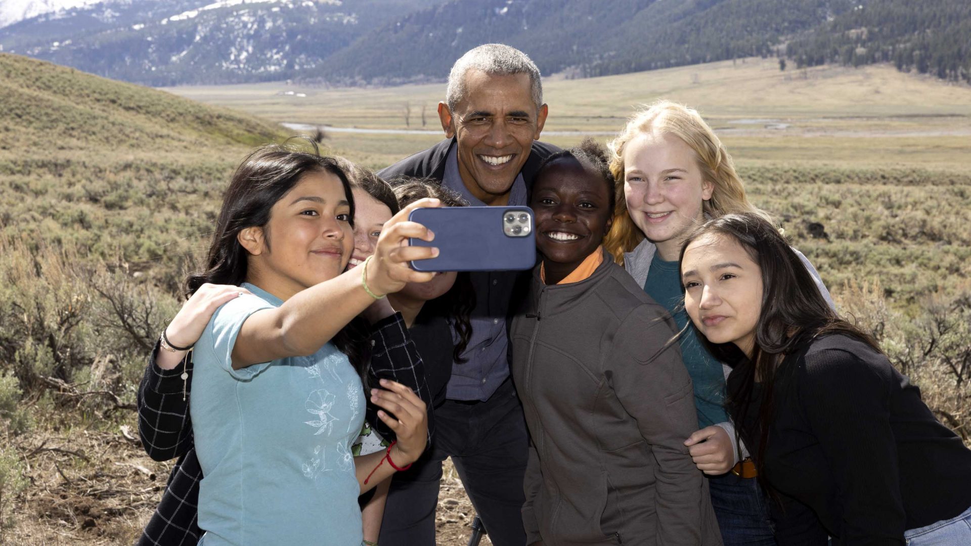 Obama smiles for a selfie with some young people while filming. He is backed by snow covered mountains.