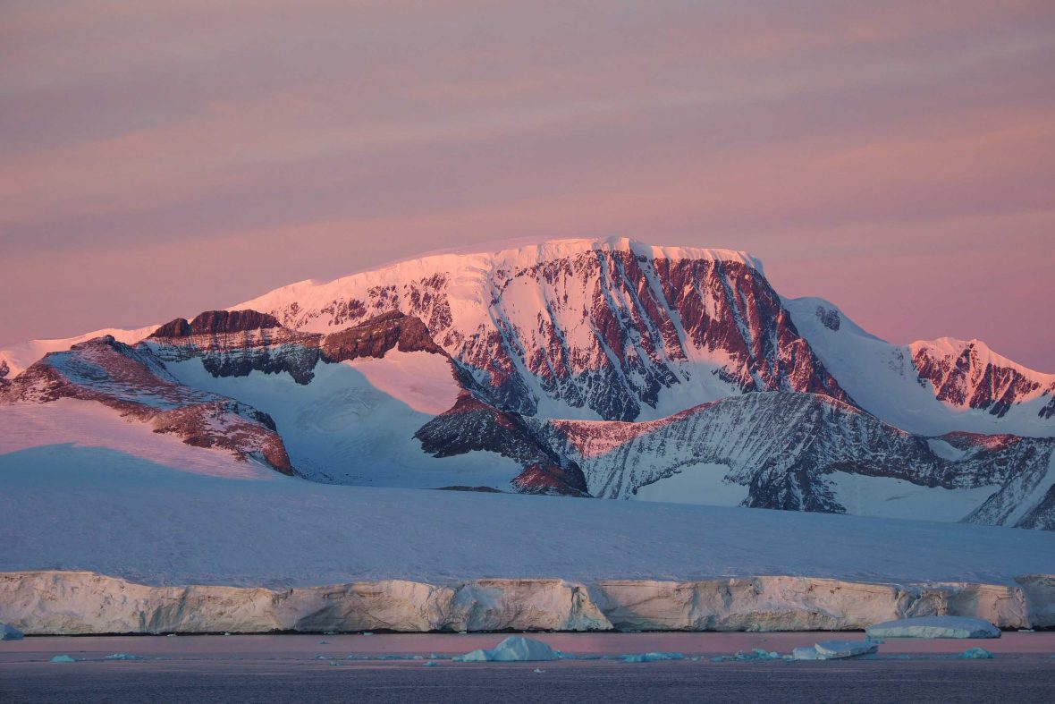 Sunrise over an iceberg. They sky is a deep pink.