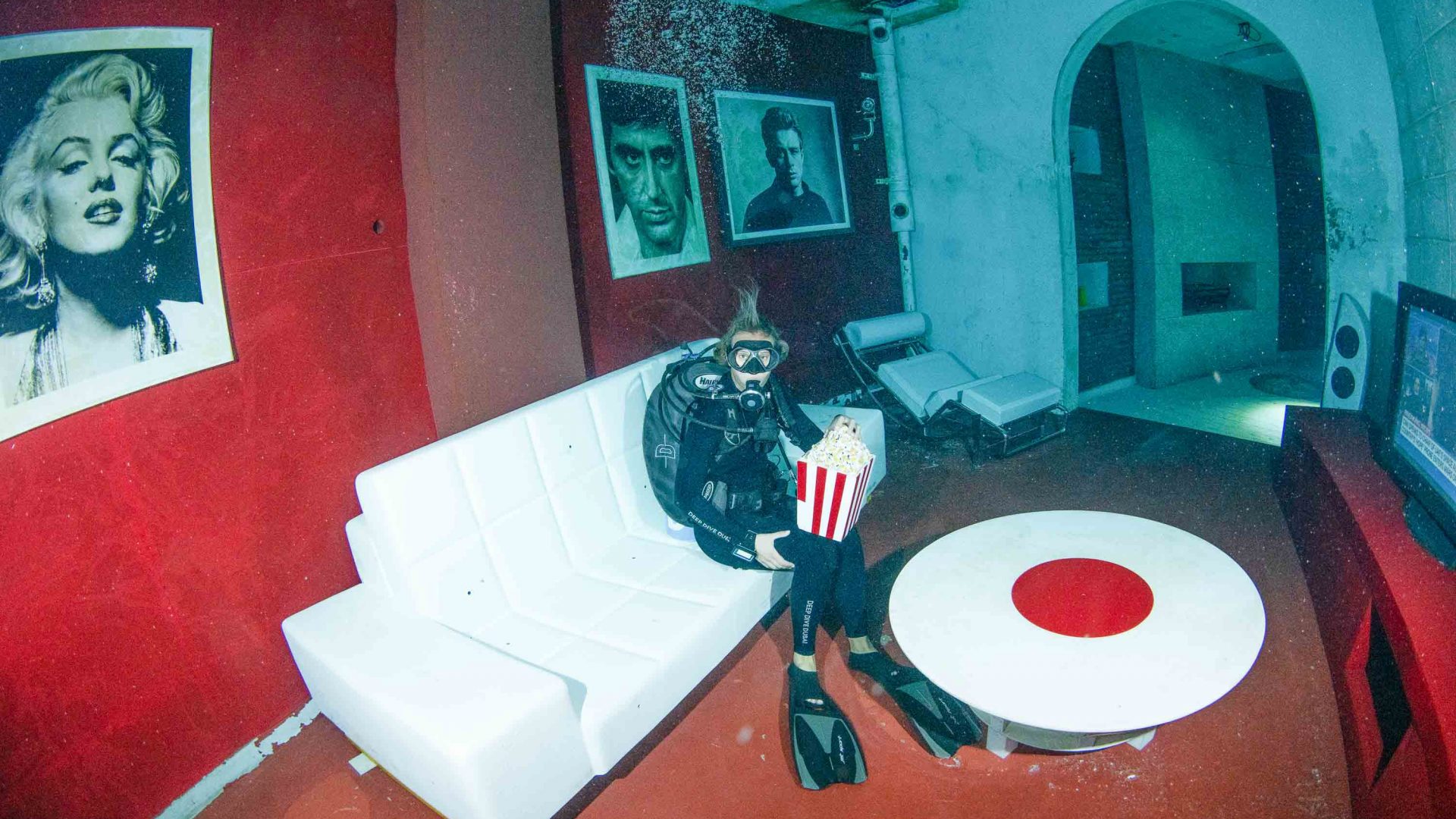 A diver holds a box of popcorn in an underwater living room.