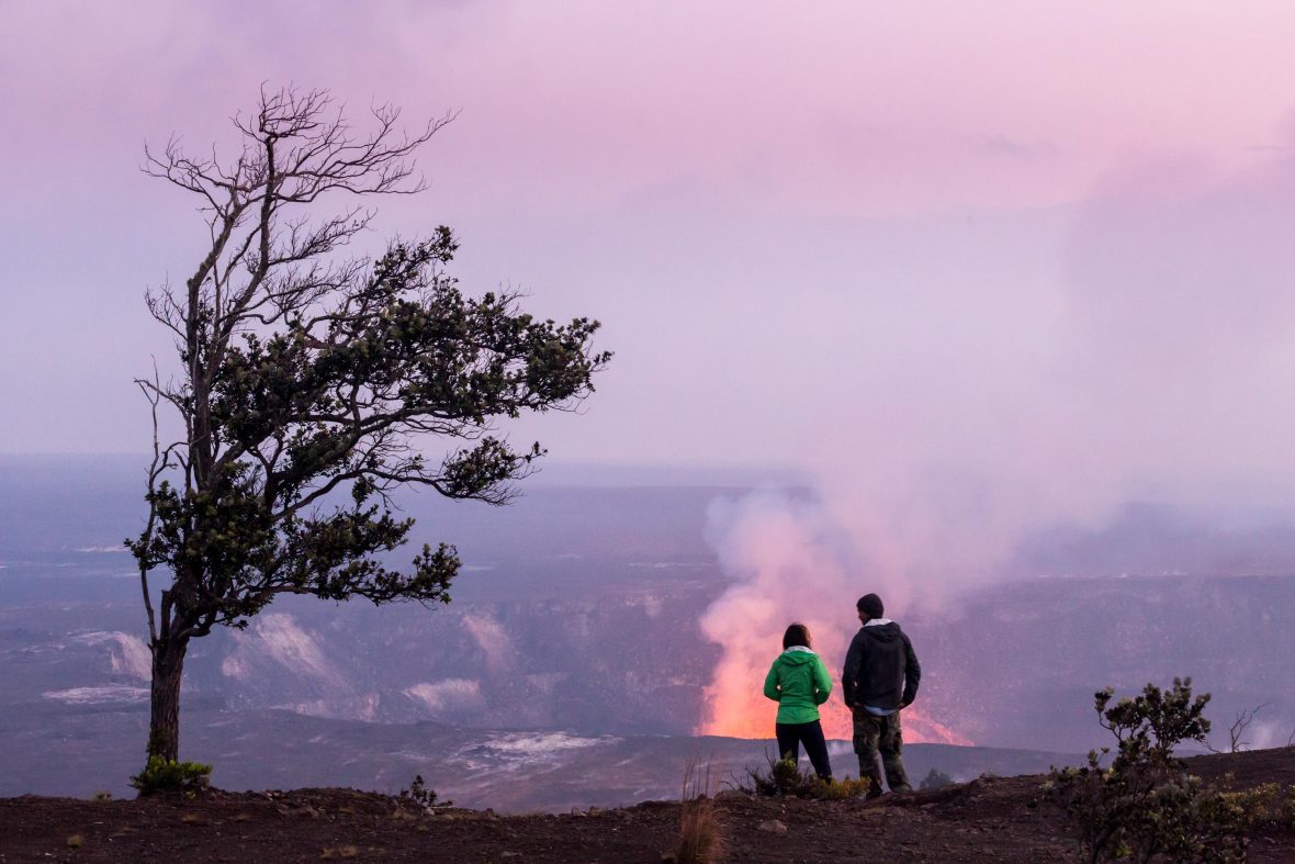 Two tourists look over the crater of a volcano that has orange lava visible..
