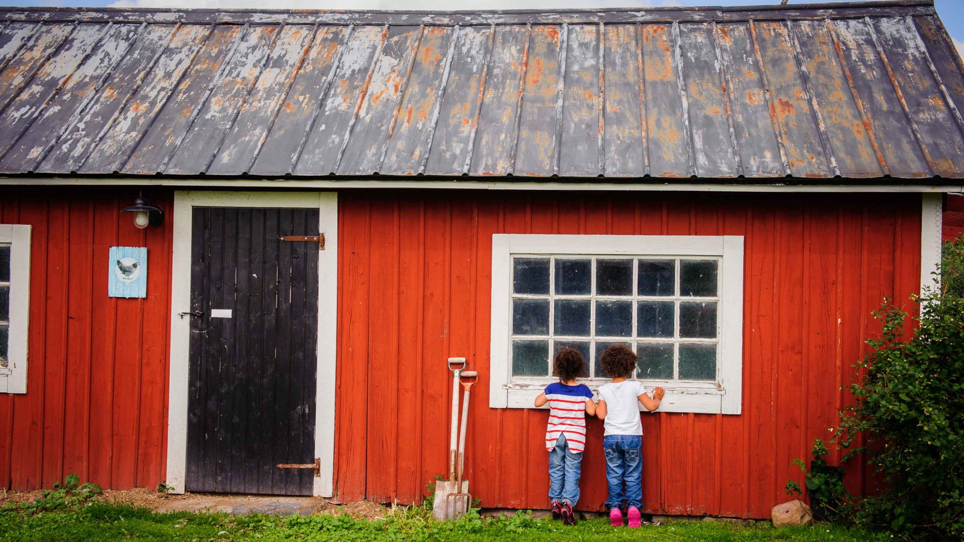 Two children look through the window of a red cottage.