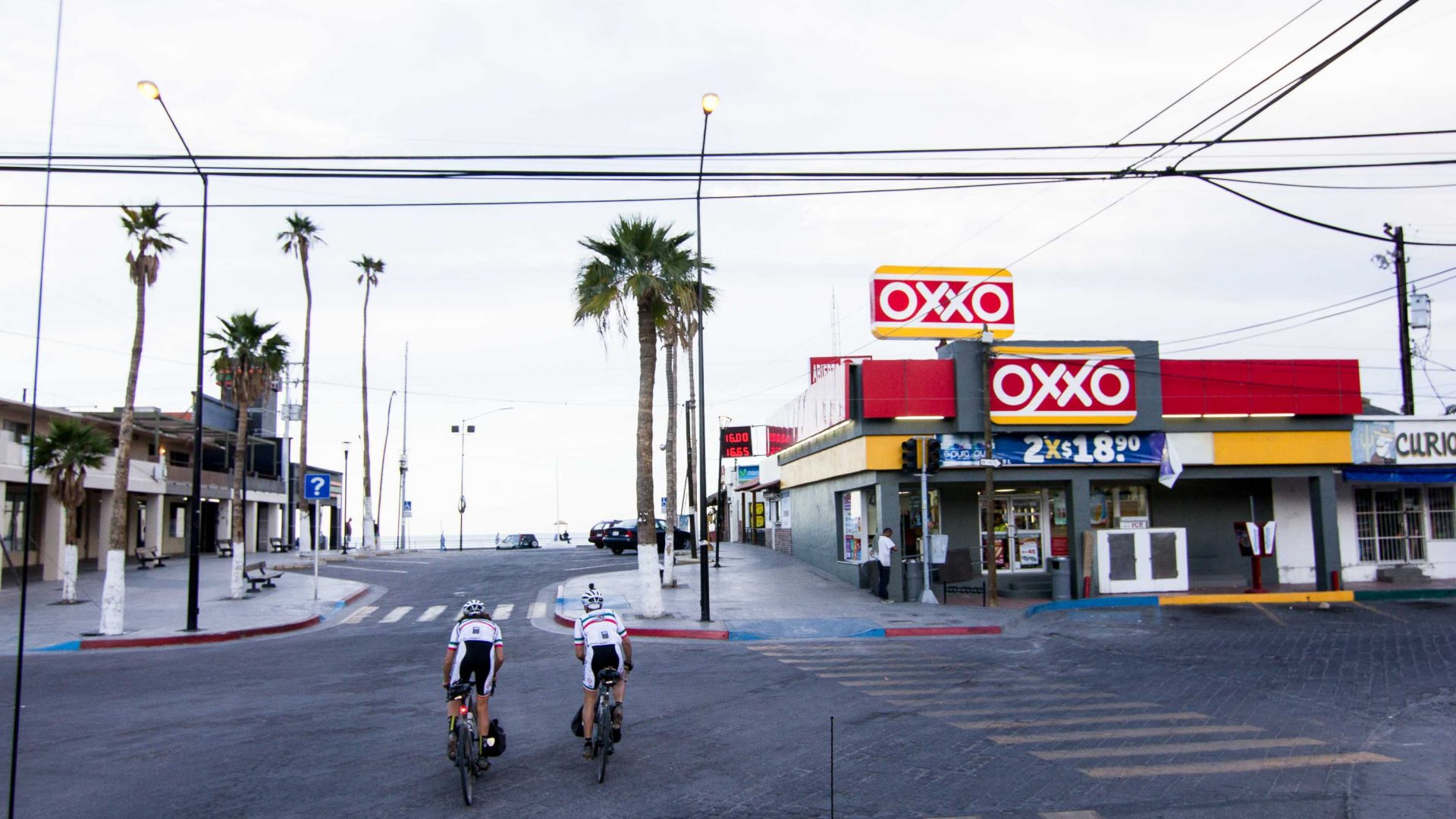 Two people cycle along the tarmac road through a town with a red and white restaurant to the right.