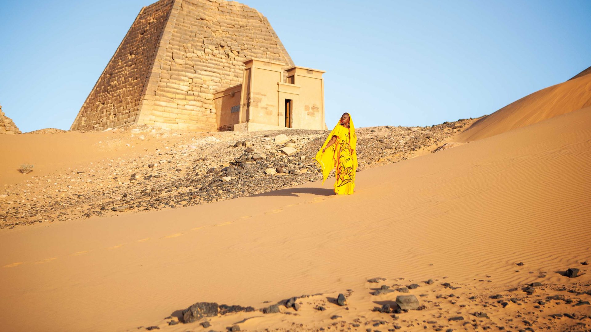 Jessica wears all yellow and stands in the desert sand with a stone structure behind her.