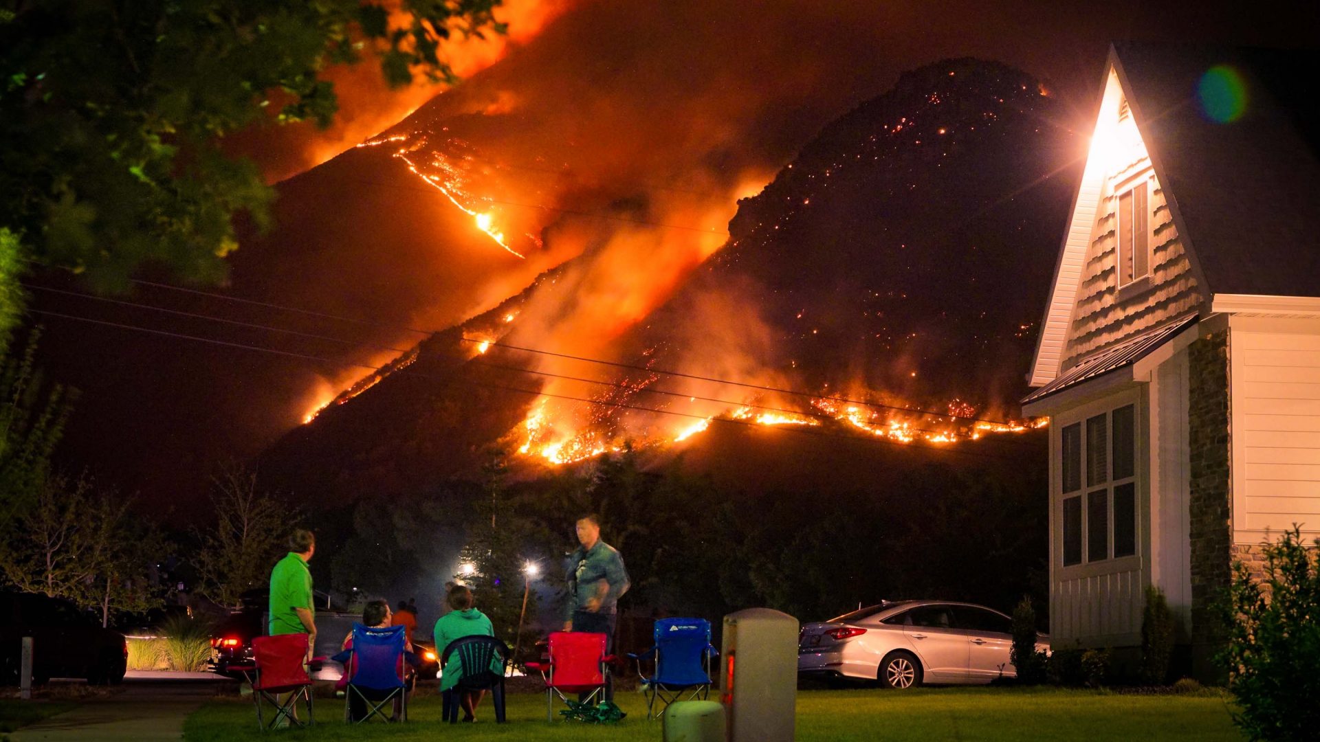 A group of people sit on chairs on the lawn of their home to watch nearby wildfires.