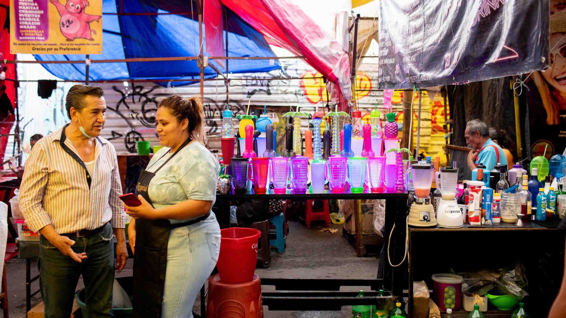A woman and man talk in front of a michelada stand selling drinks in penis shaped cups.