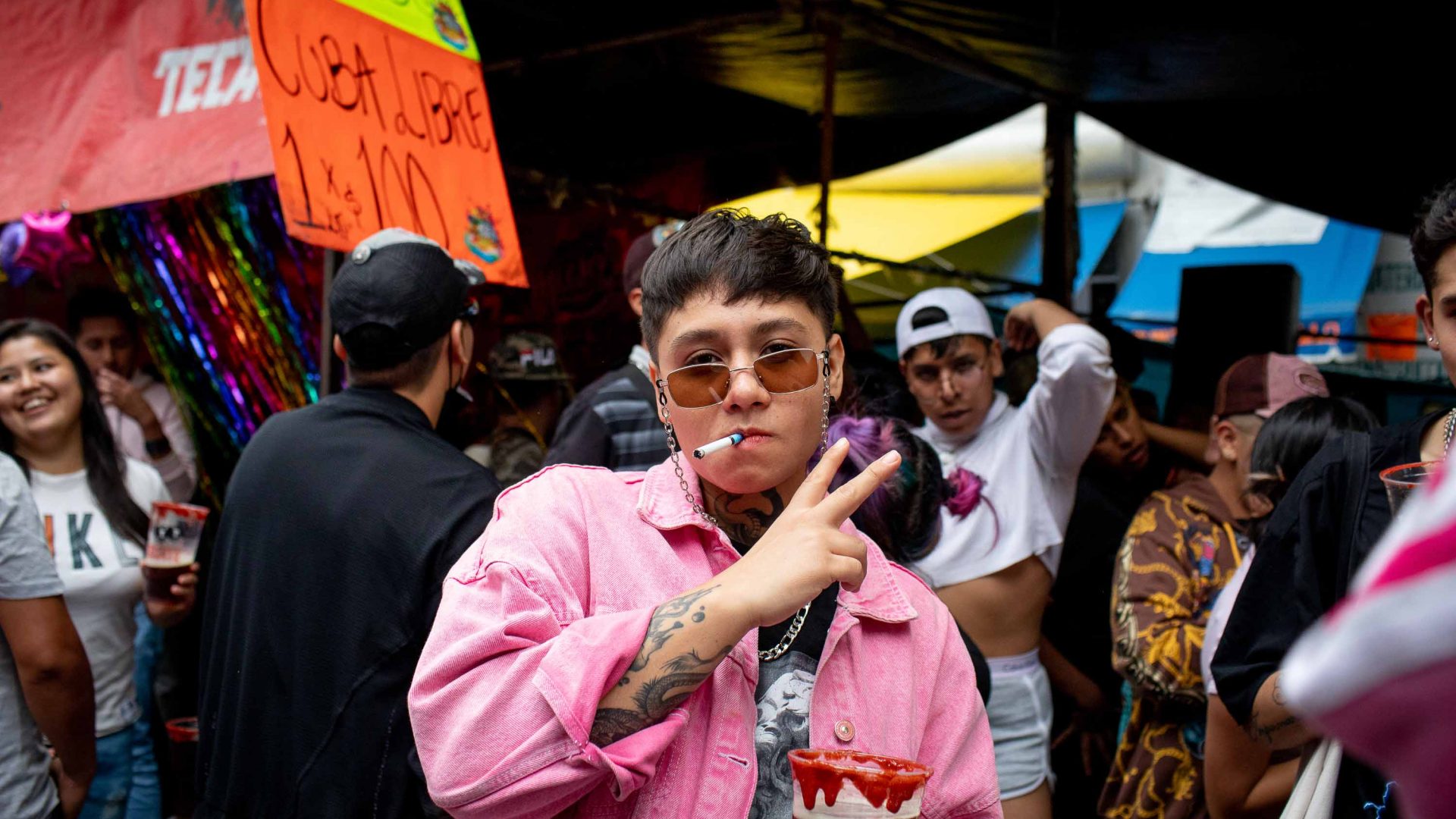 A man poses for a photo, michelada in hand. He is wearing sunglasses and smoking a cigarette.