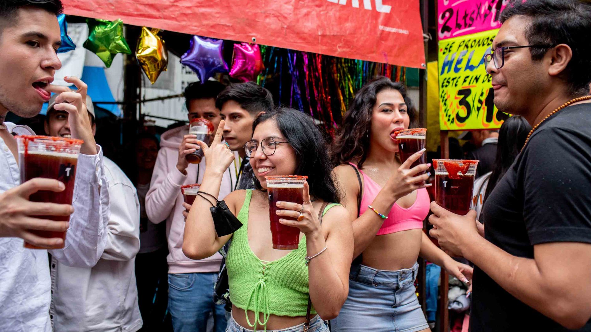 Partygoers drink micheladas under a red sign.