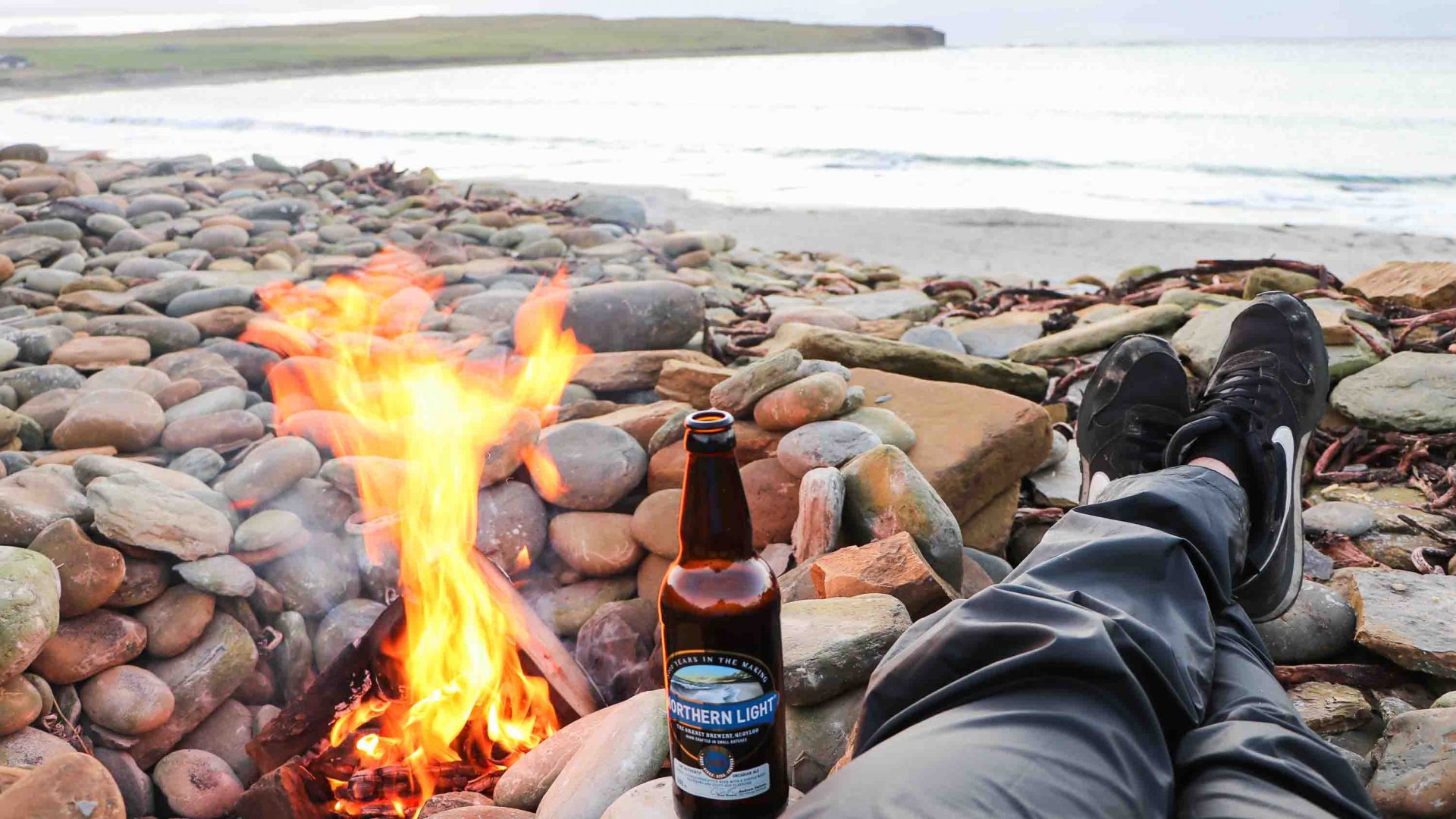 A beer bottle resting on legs alongside a small fire, with views of a lake.