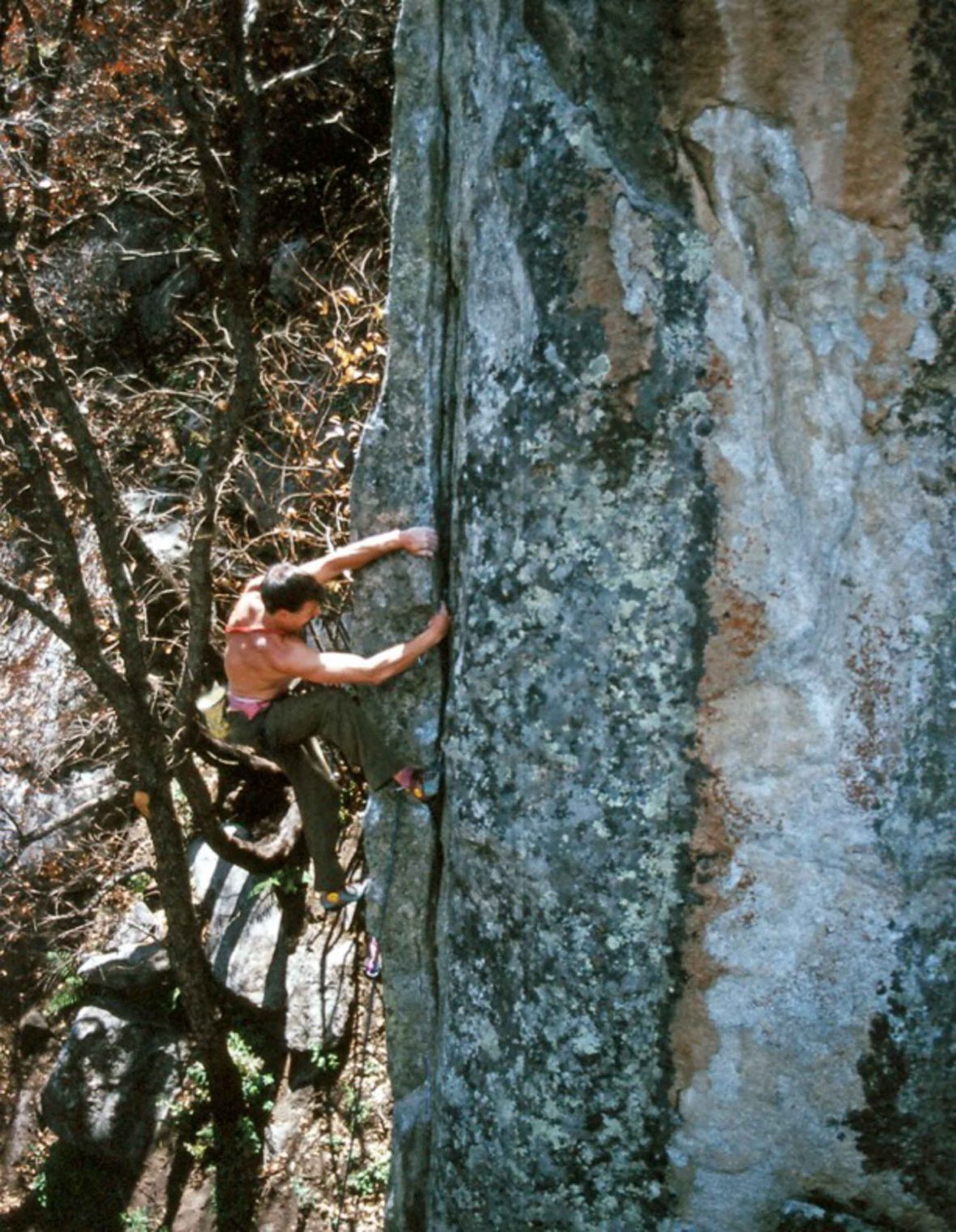 A young Ken Yager with no shirt on climbs a rock face.