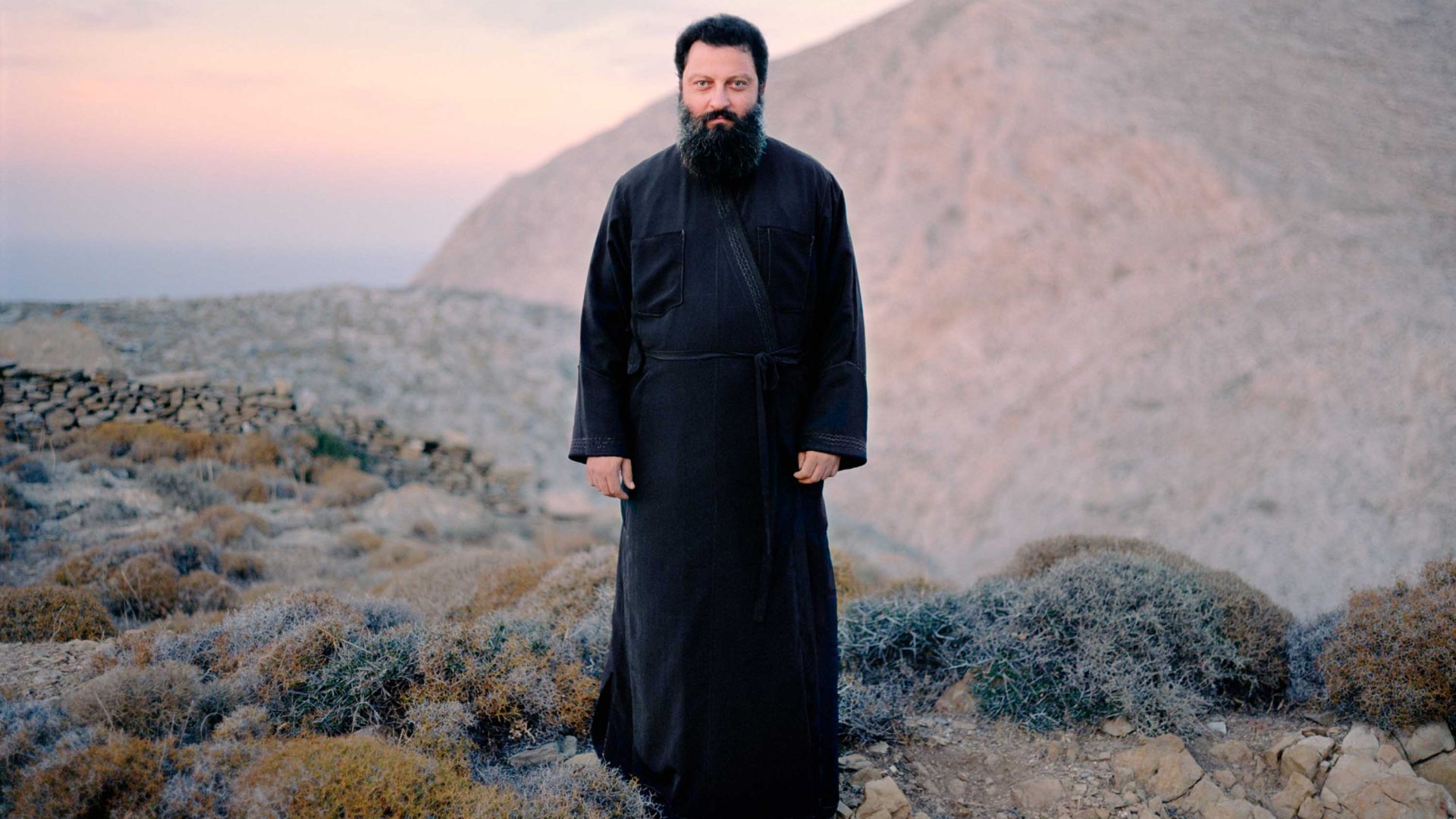 A man in a black robe in an arid landscape looks straight to camera.