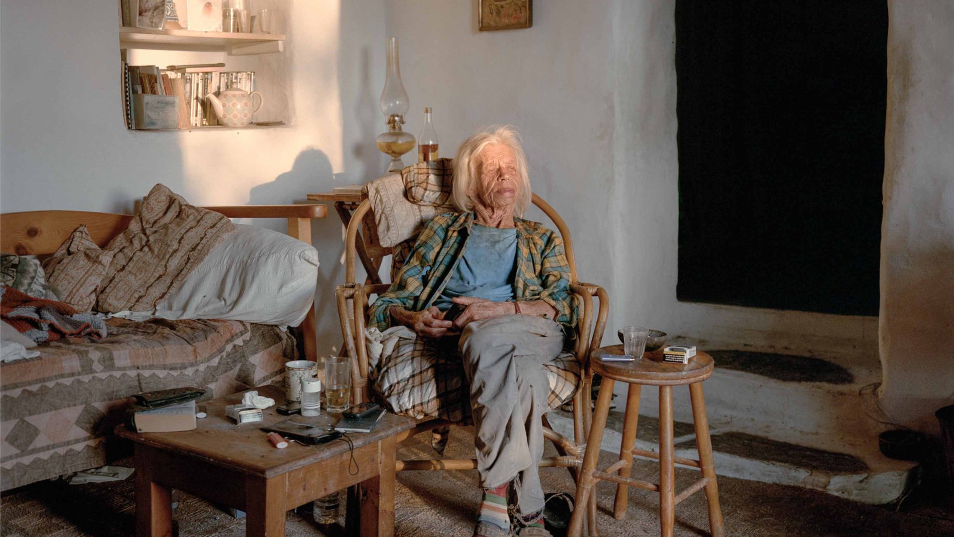 An elderly lady sits in a chair in her home. Soft light falls on her face.