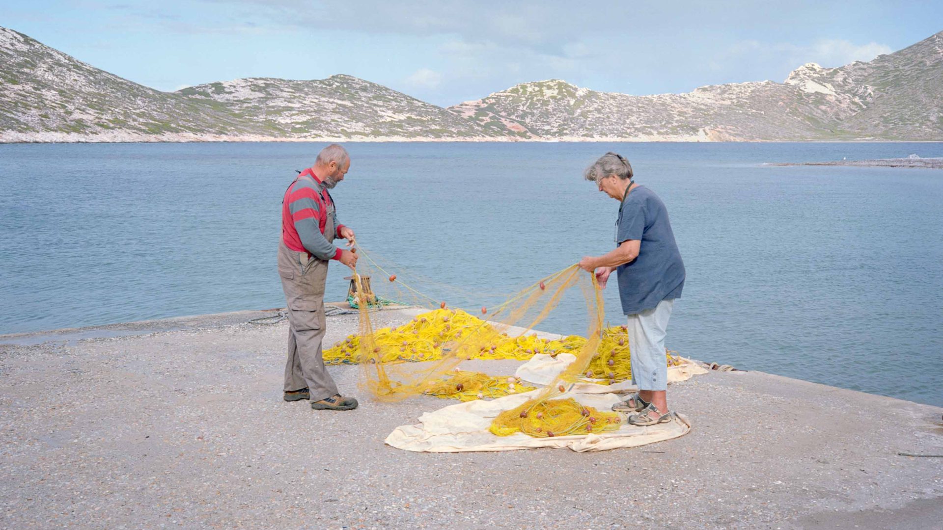 Two people work to untangle a fishing net by the sea.