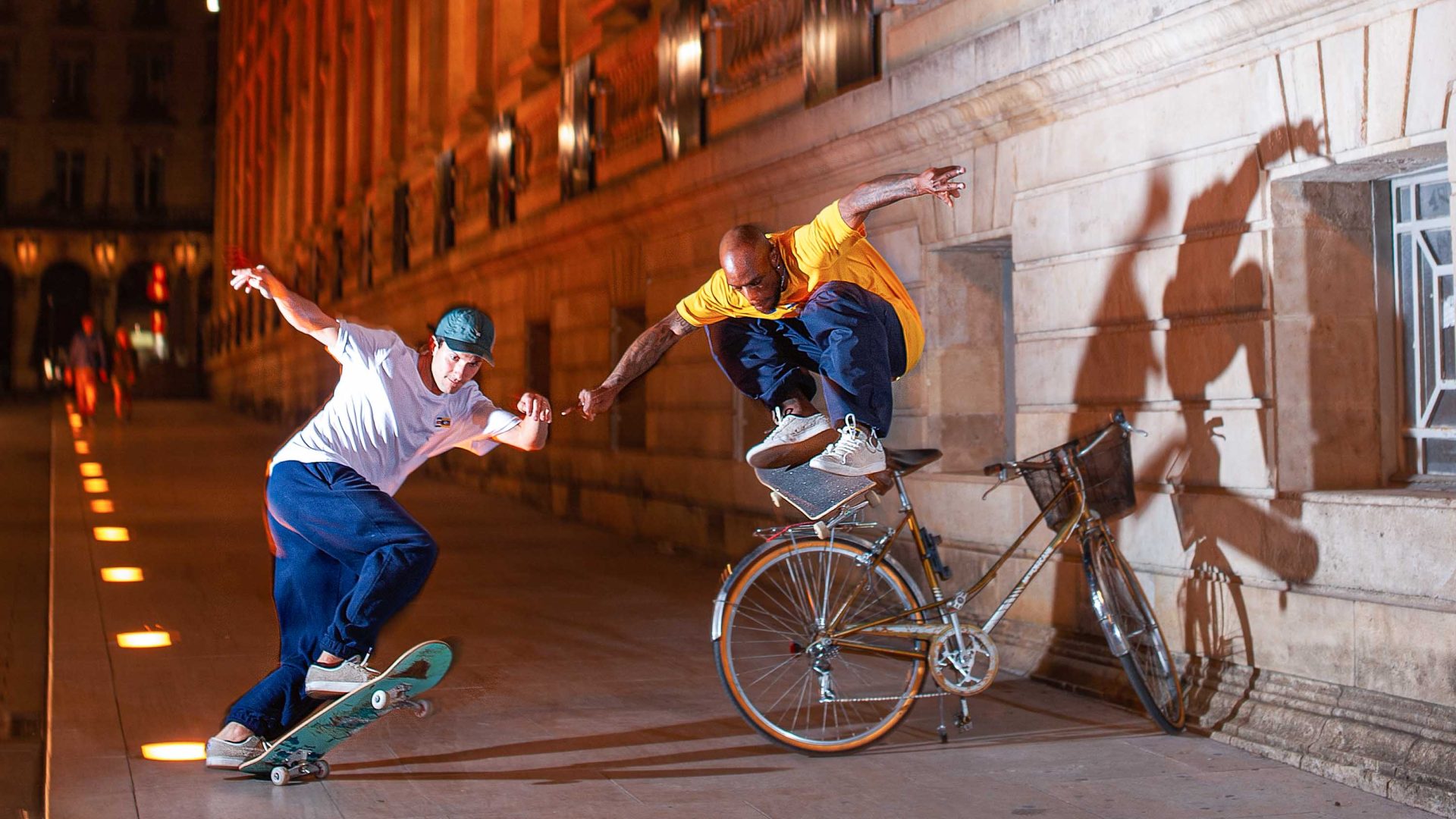 Leo on his skateboard while another man does an ollie over a bike.