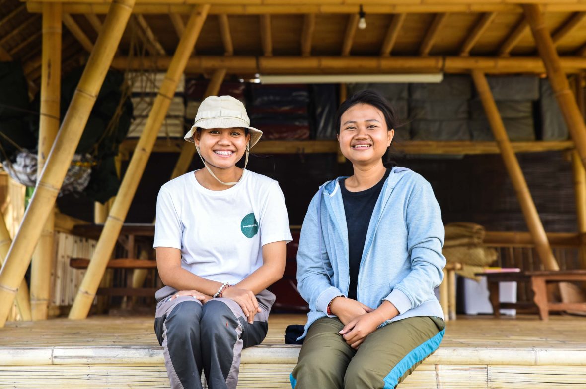 Two women smile on the steps of a bamboo hut