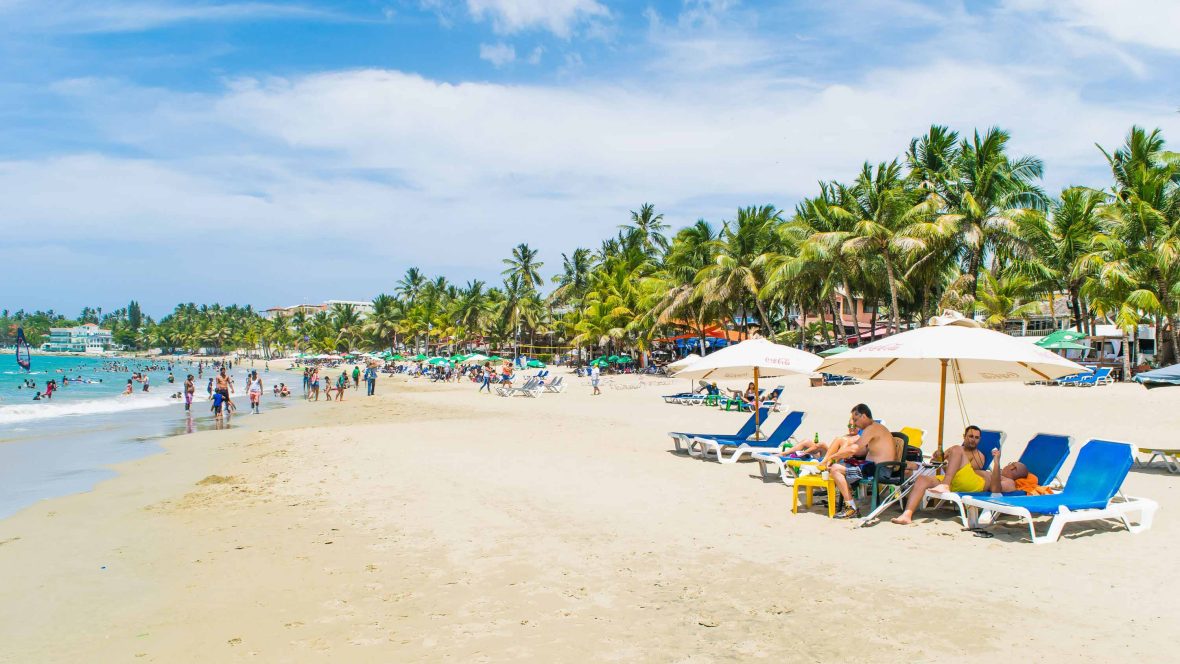 Tourists line a beach with umbrellas and palms.