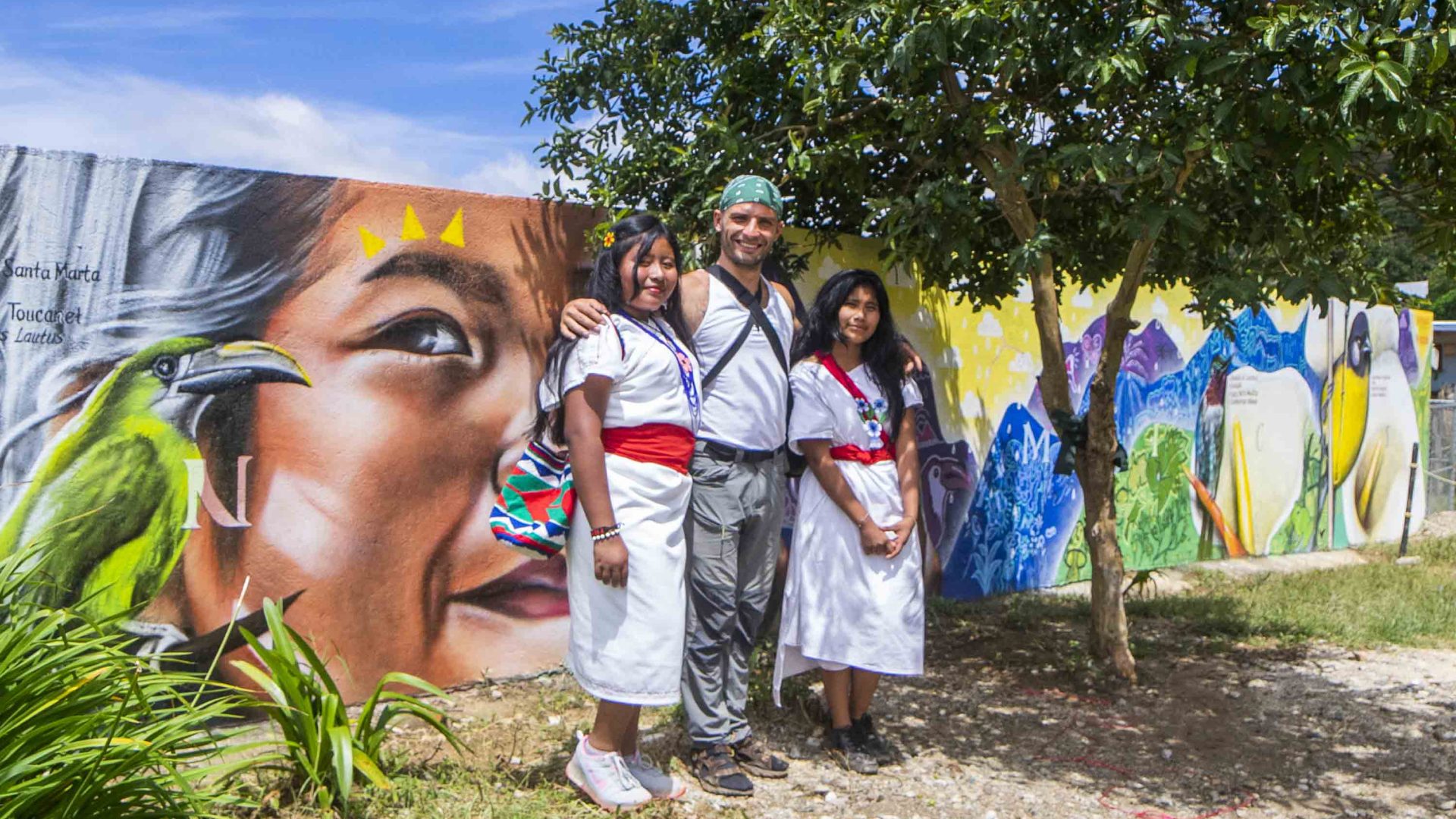 Two local indigenous women stand with a man in front of a painted mural.