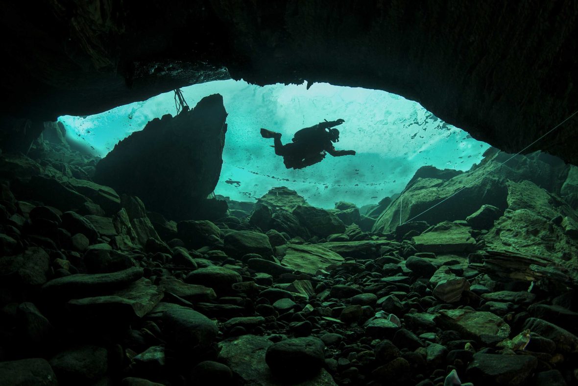 A diver swims past a cave with green water.