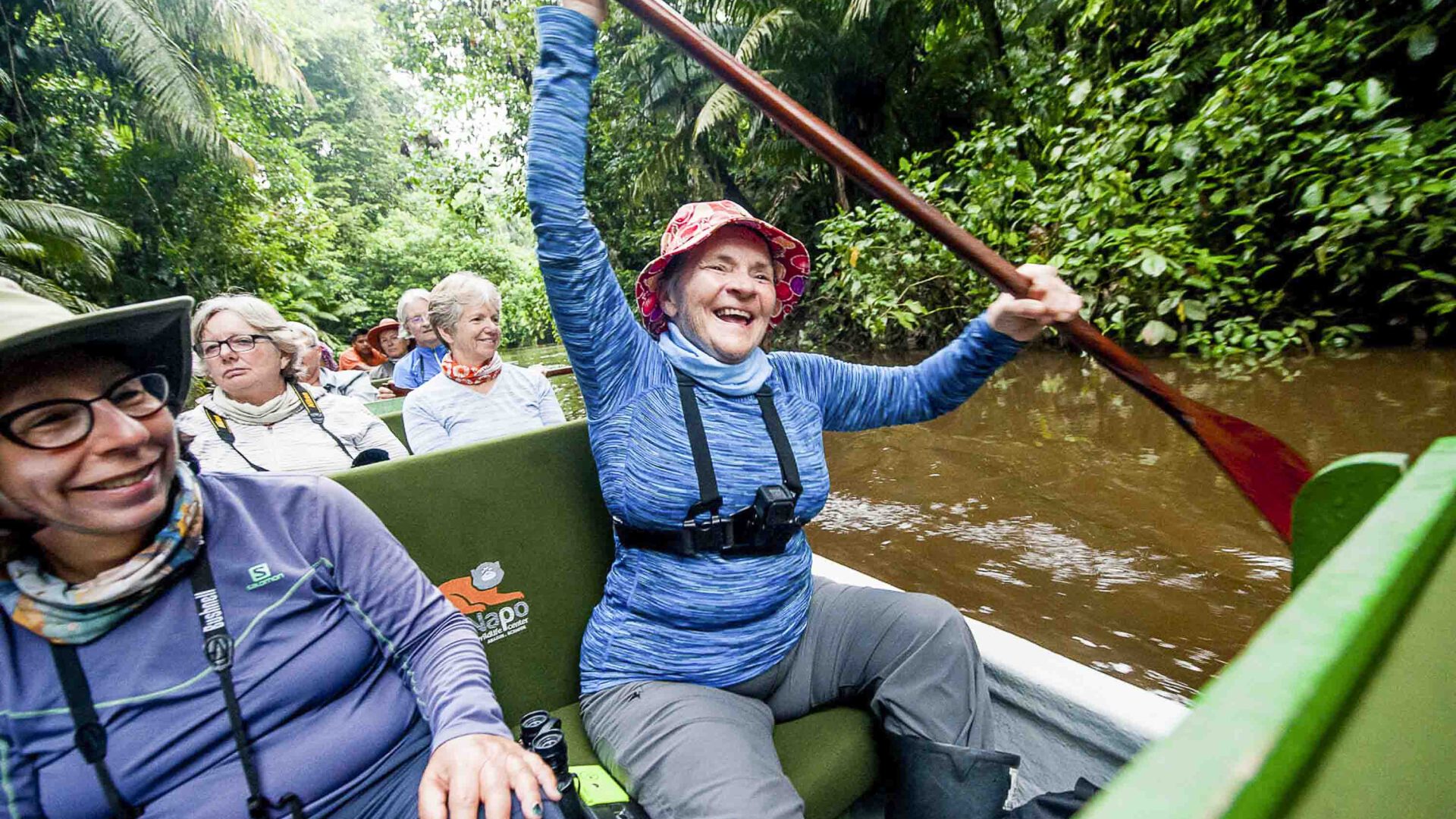 Women paddle down a river in a boat. One of them is laughing.
