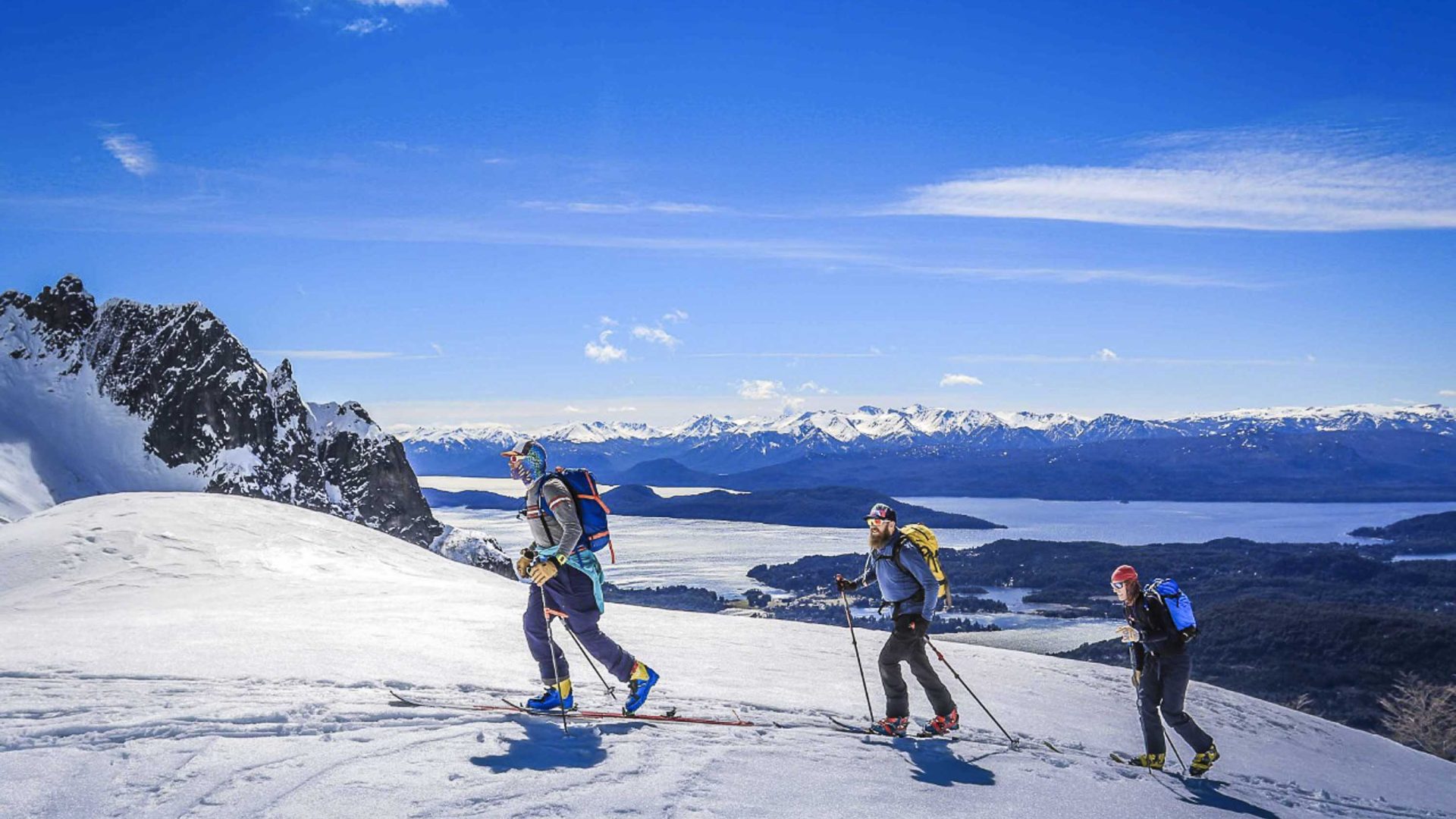 Three skiiers ascend a mountain with a lake behind them.