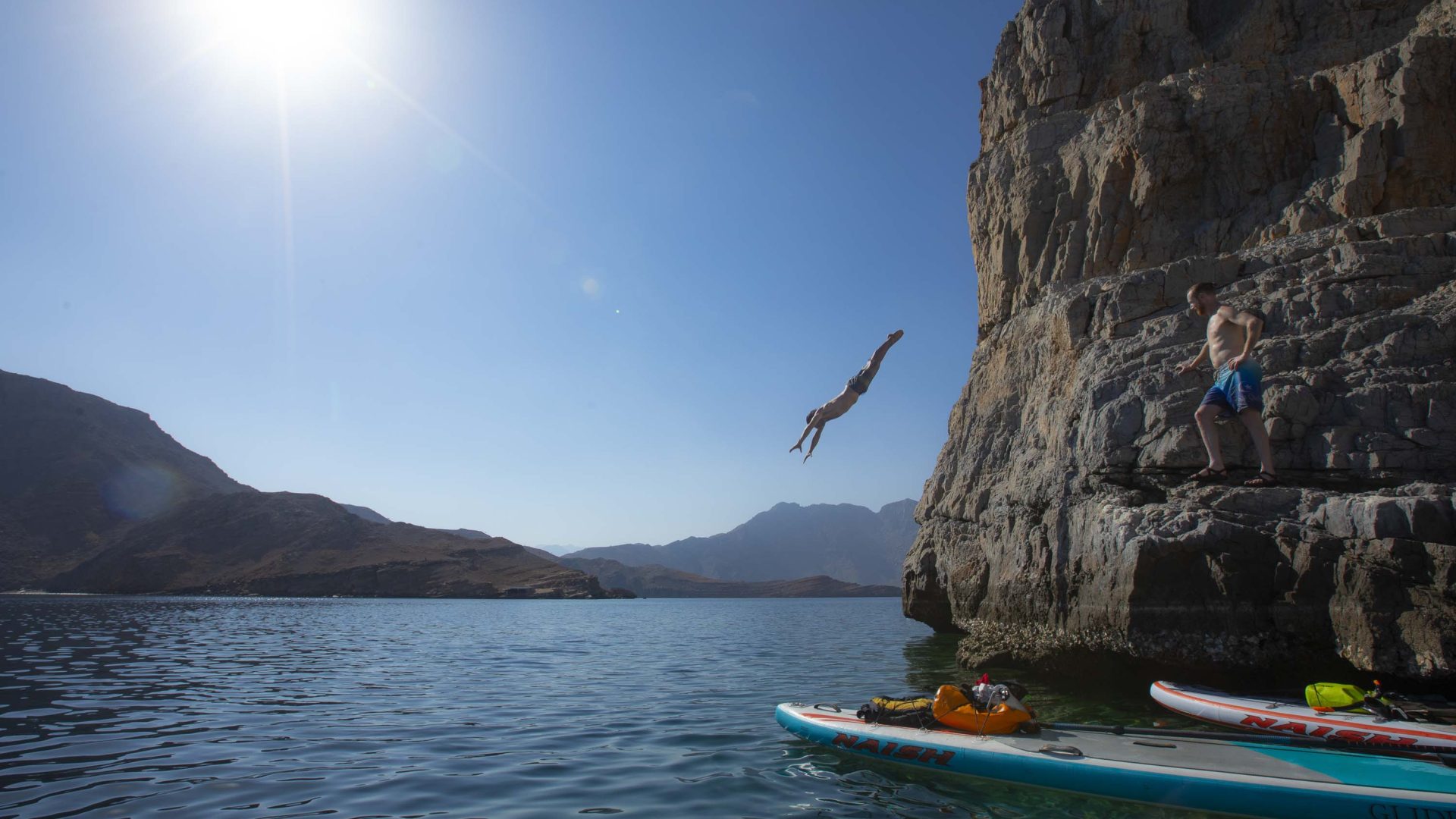 A person jumps from a rock face while paddle boards are seen below in the foreground.