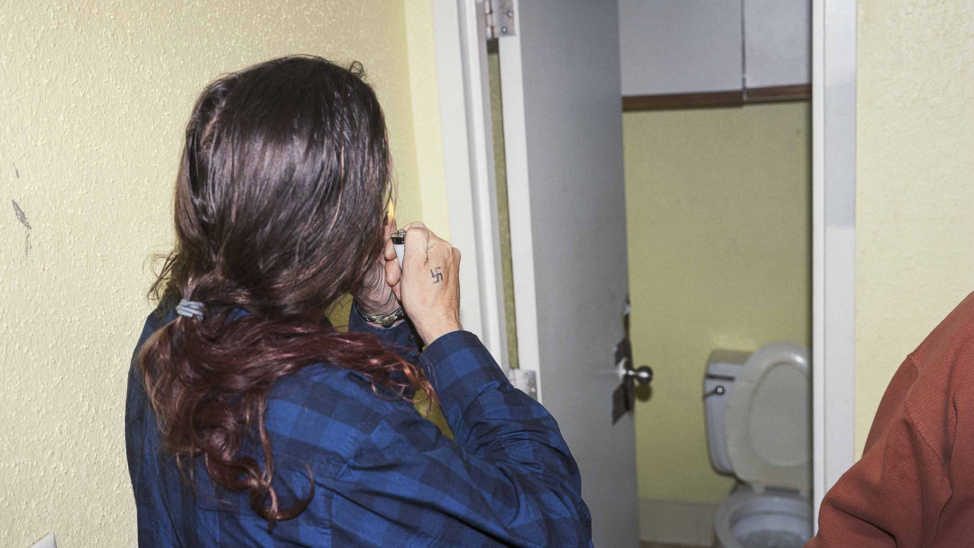 A man with long hair smokes in front of a toilet door.