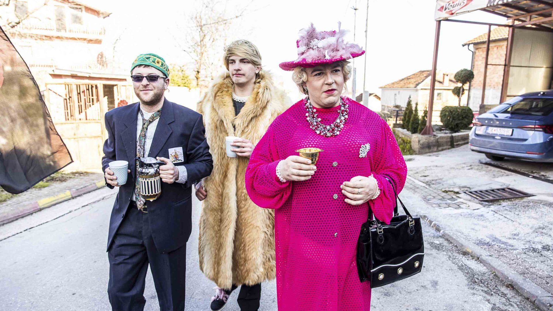 Three people walk down the street, one dressed as the Queen.