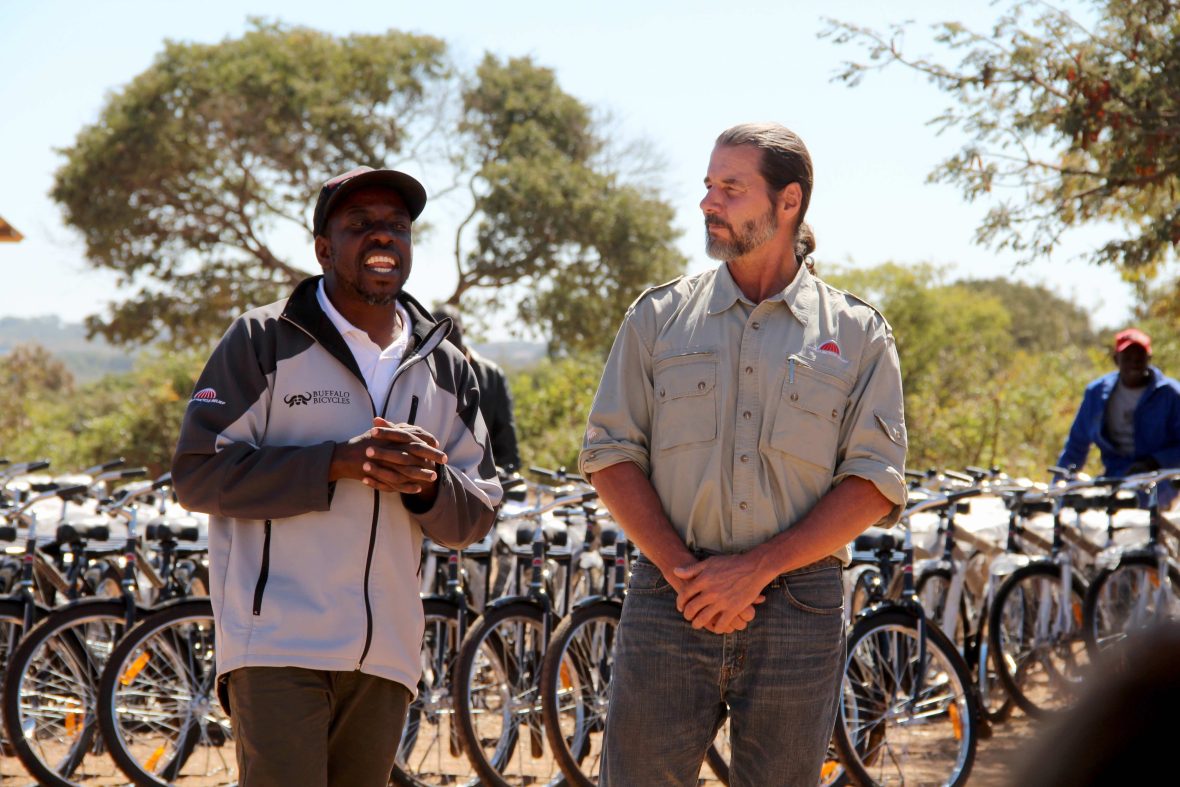 Two men talking and smiling with a row of bikes behind them.