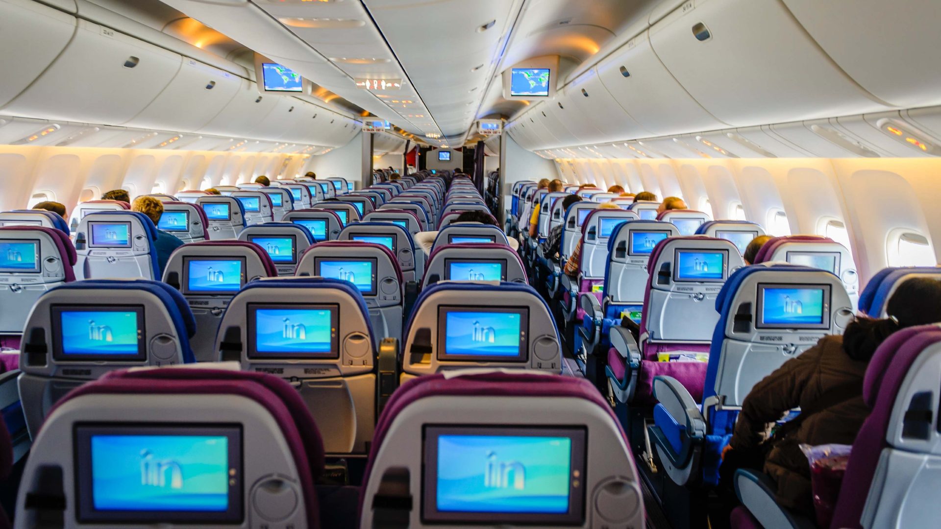 Looking down the aisle of a plane showing rows of seats and TV screens on.