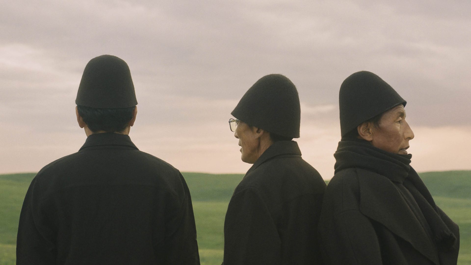Three men in black robes and hats stand in a field looking away.