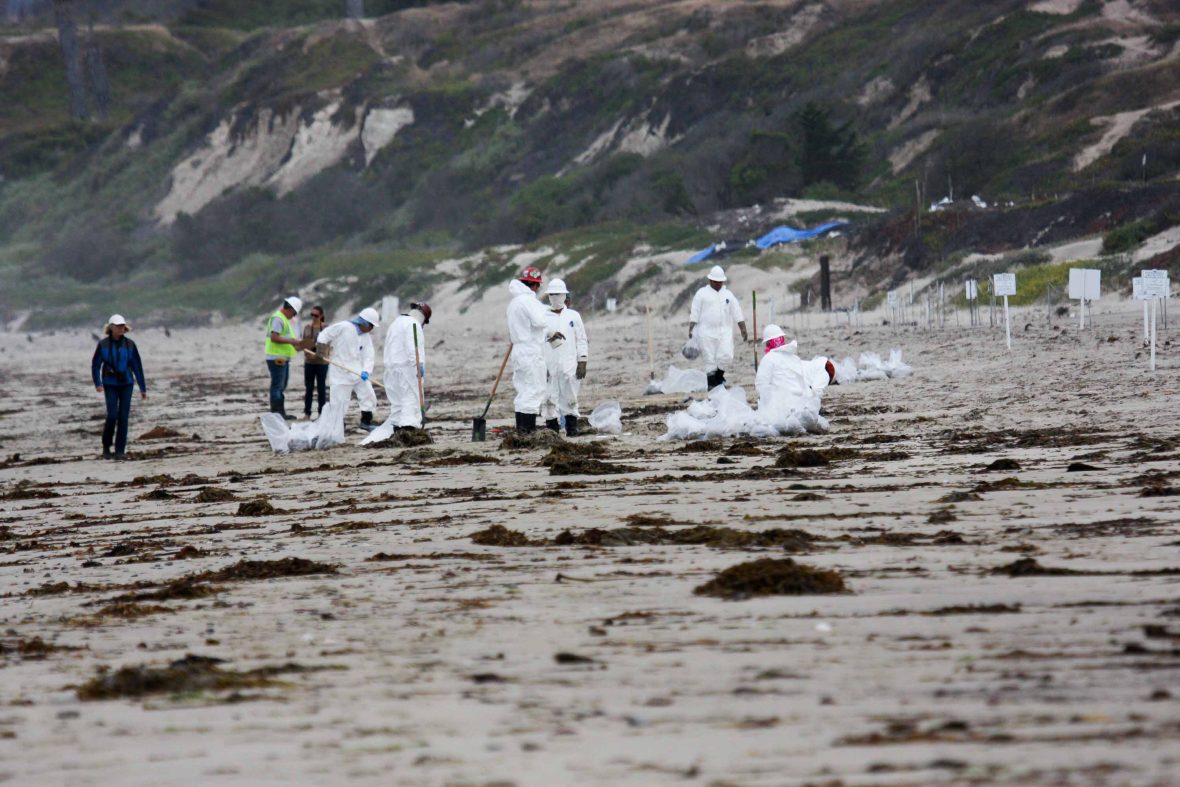 People in white protective gear clean up the oil and other mess from a beach.